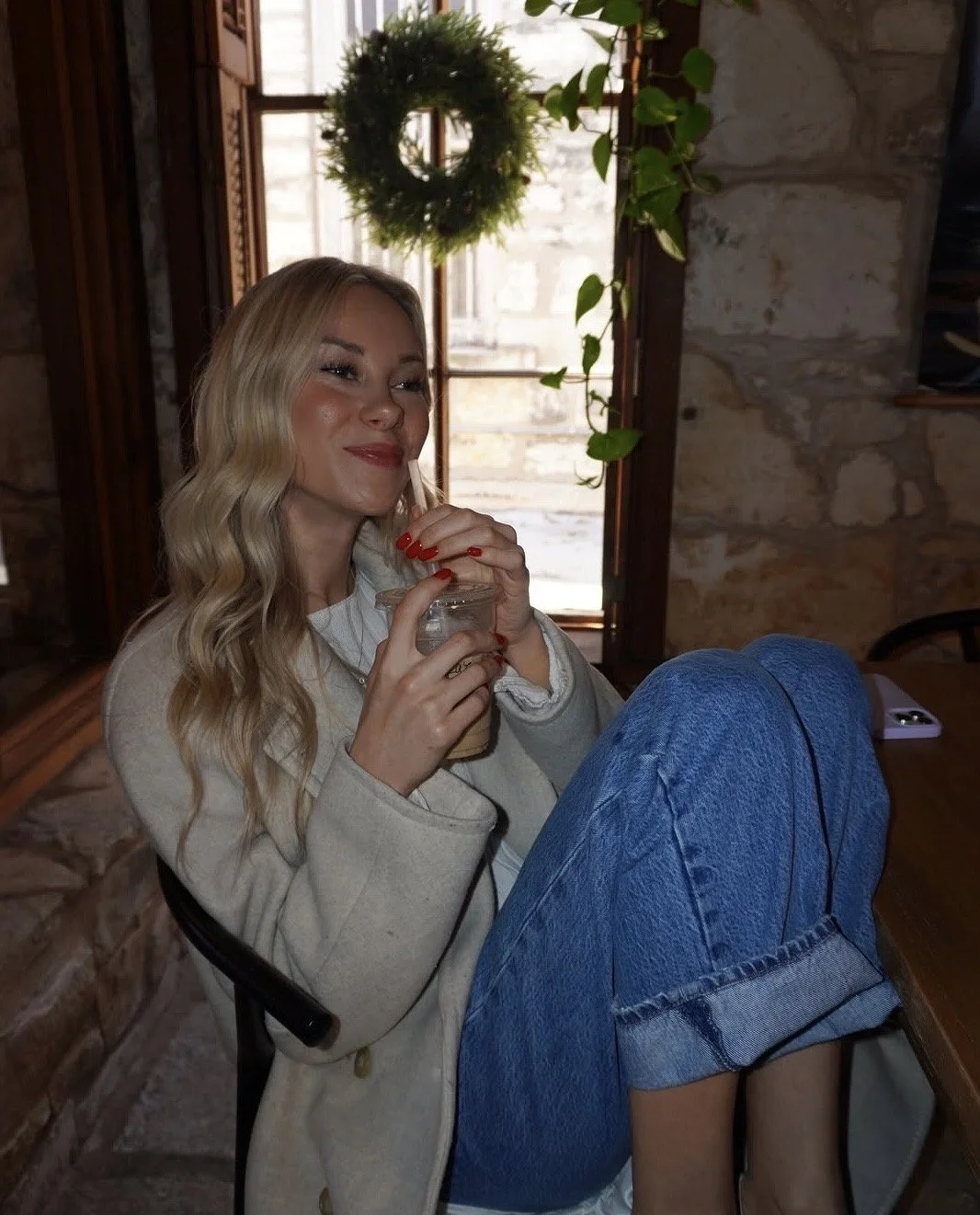 A woman with long blonde hair, red manicure, and a beige coat, sitting and holding a drink in a glass jar with a straw, inside a cozy room with stone walls and a window with a door and green plant in the background.