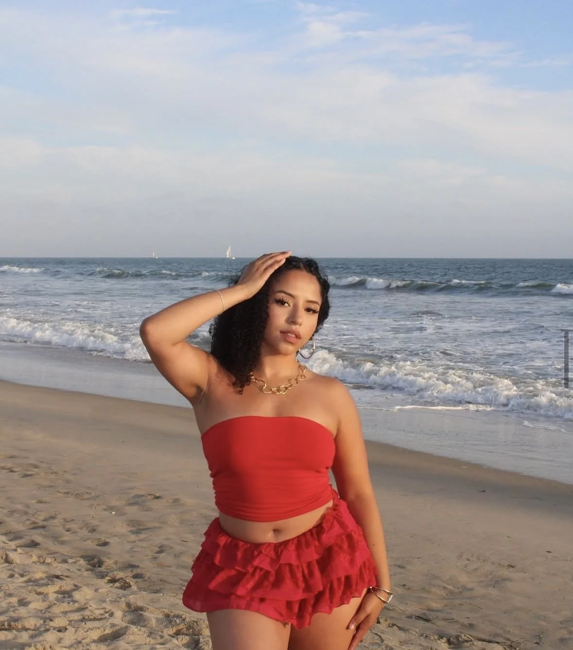 A woman in a red tube top and matching ruffle shorts standing on a sandy beach with the ocean and sailboats in the background.