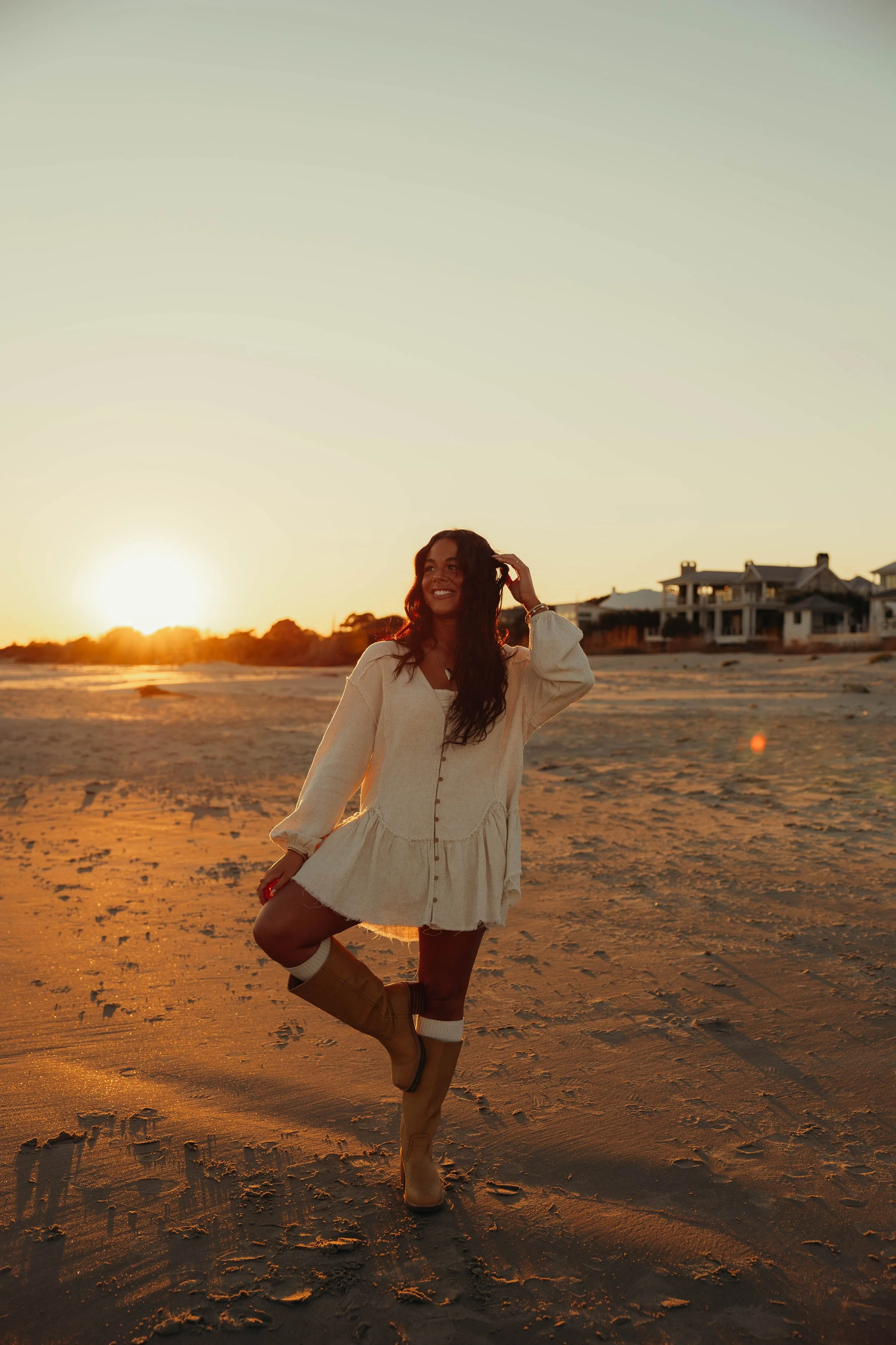 Woman in white dress and brown boots standing on sand at sunset, smiling and touching her hair, with beach houses in the background. Kailey Jeffcoat 