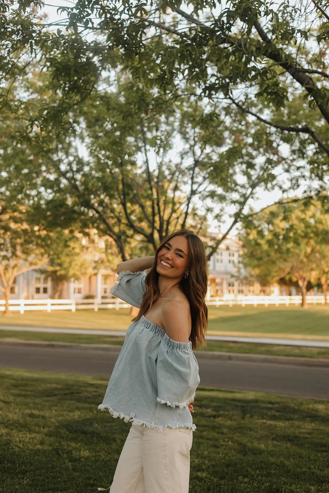 A young woman with long brown hair and a bright smile standing outdoors in a park with trees and houses in the background during golden hour. She is wearing a light off-shoulder top and cream-colored pants, with one hand behind her head.