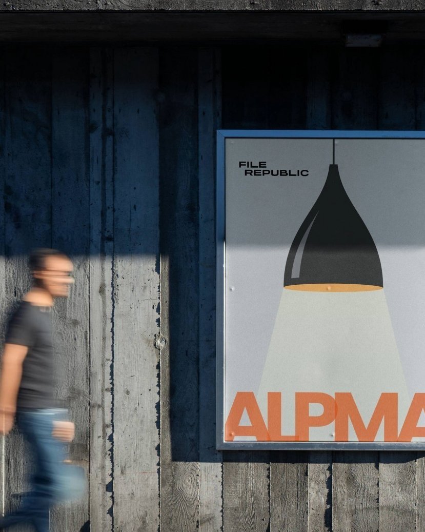 A blurred person walks past a poster with a lamp illustration and the words "File Republic" and "Alpma" on a wooden wall.
