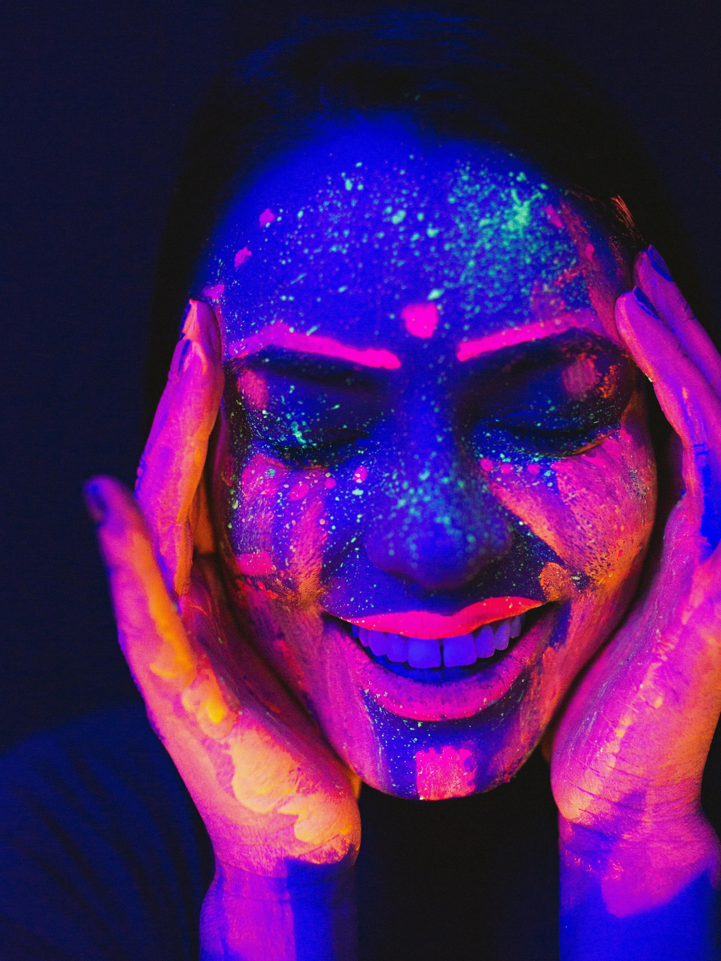 Person with face painted in neon colors, smiling, with hands near face, under black light revealing glowing paint.