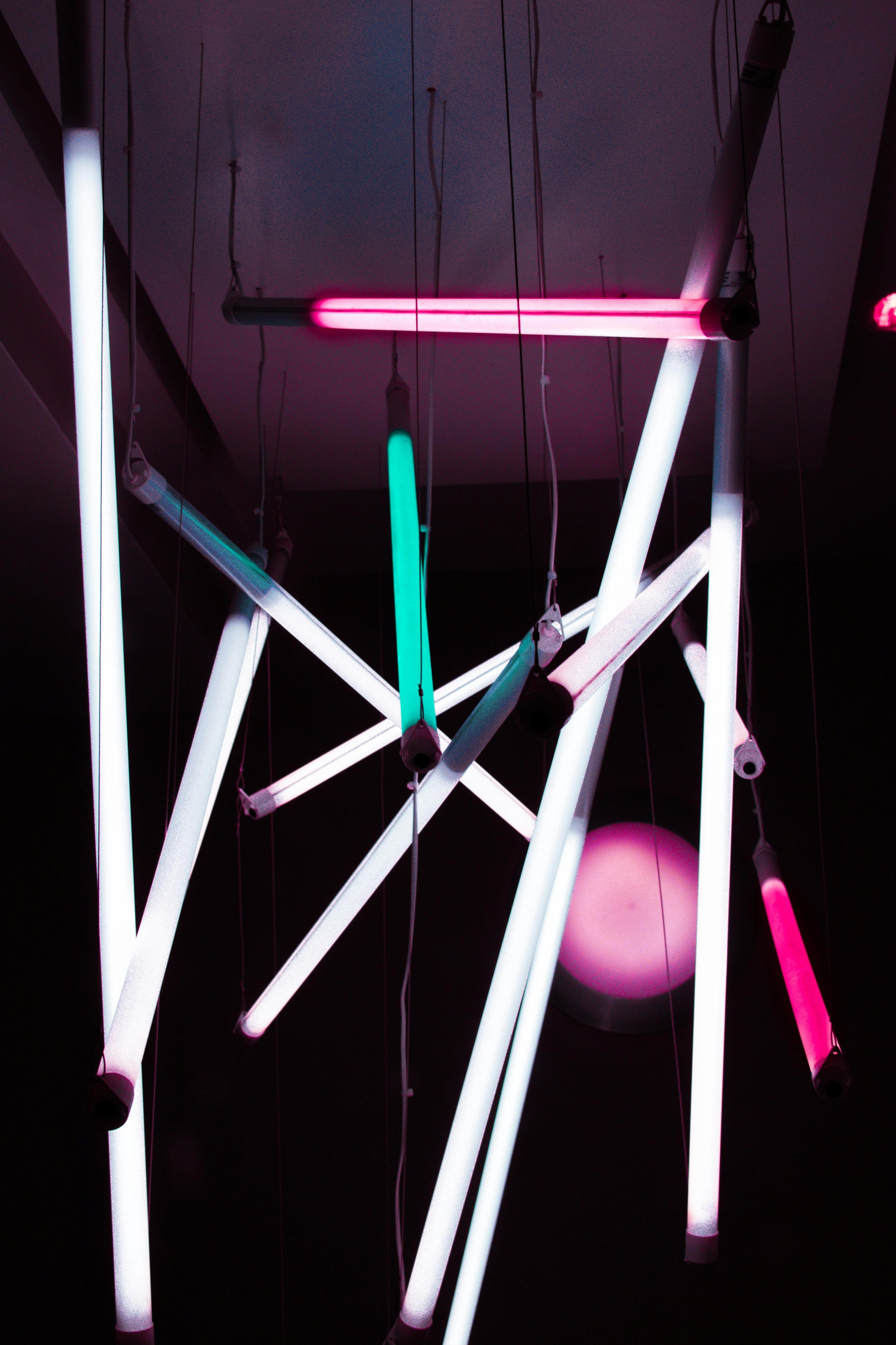 A modern art installation made of glowing neon light tubes in pink, white, and green colors hanging from the ceiling against a dark background.