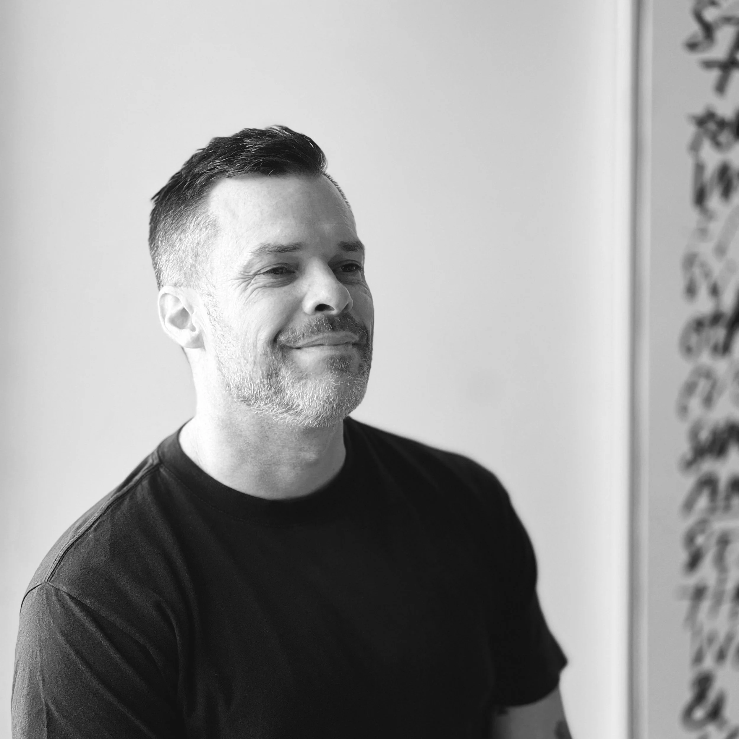 Black and white photo of a smiling man with short hair and a beard, wearing a black t-shirt, standing indoors against a plain wall.