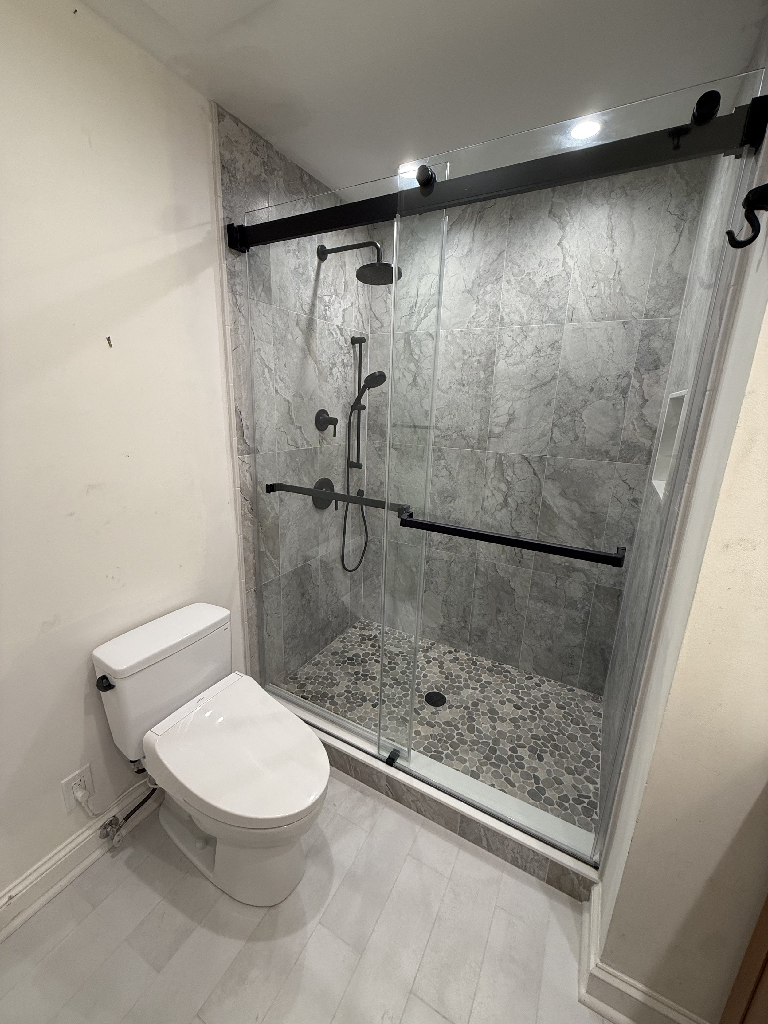 A modern bathroom with a white toilet and a glass-enclosed shower with gray marble tiles and pebble floor.