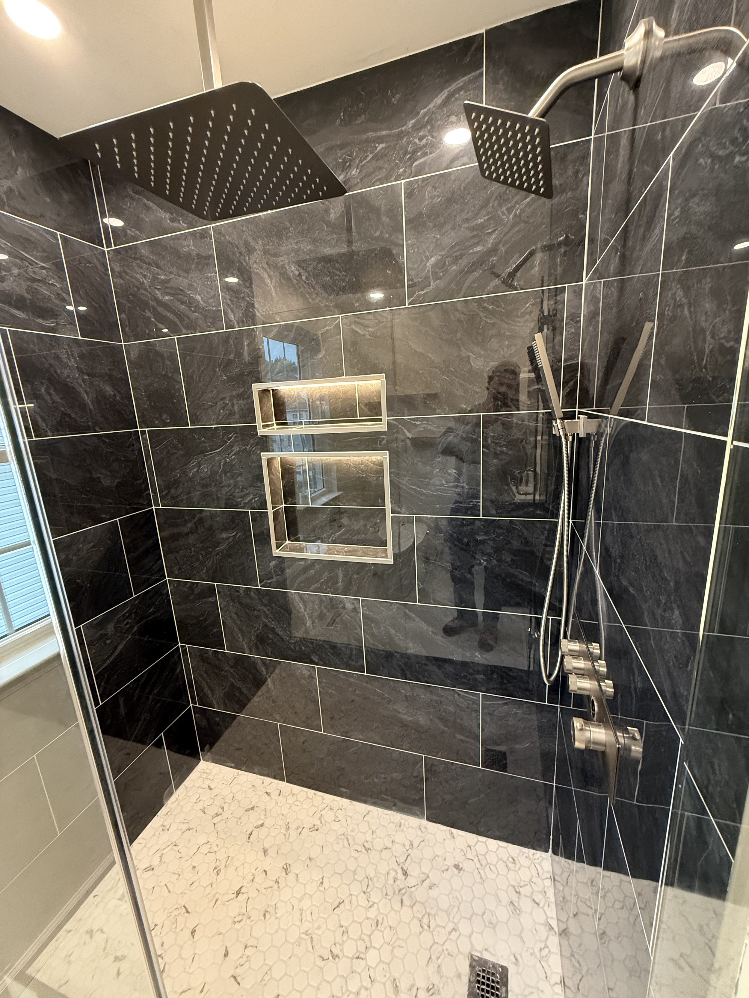 A modern shower area with large black marble tiles on the walls, a black rain shower head mounted from the ceiling, a handheld shower with a flexible hose, and two built-in wall niches with lighting. The floor has small white hexagonal tiles, and there is a floor drain.