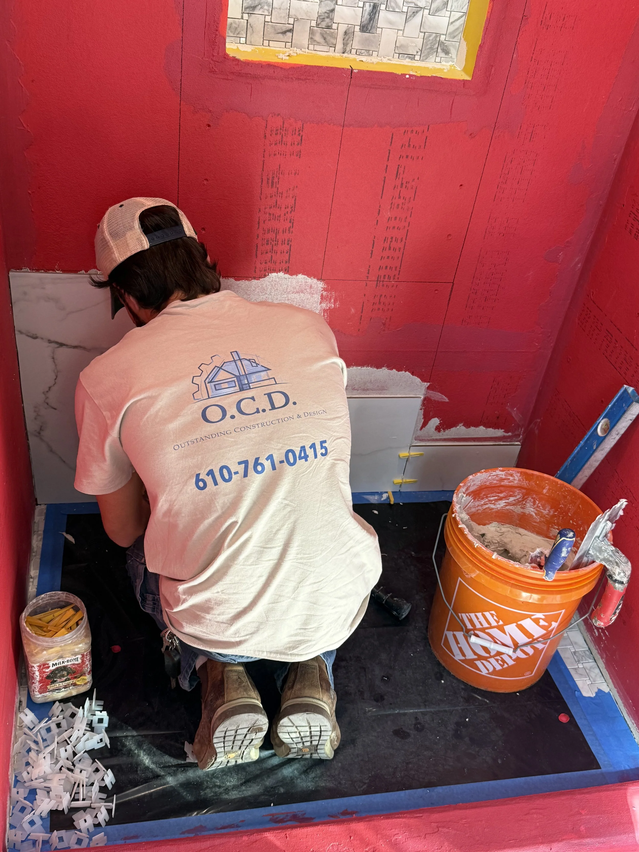 Worker with back facing, wearing a beige baseball cap and tan boots, installing ceramic tiles on a red wall in a bathroom under construction. Tools and supplies, including an orange Home Depot bucket filled with mortar, are nearby.