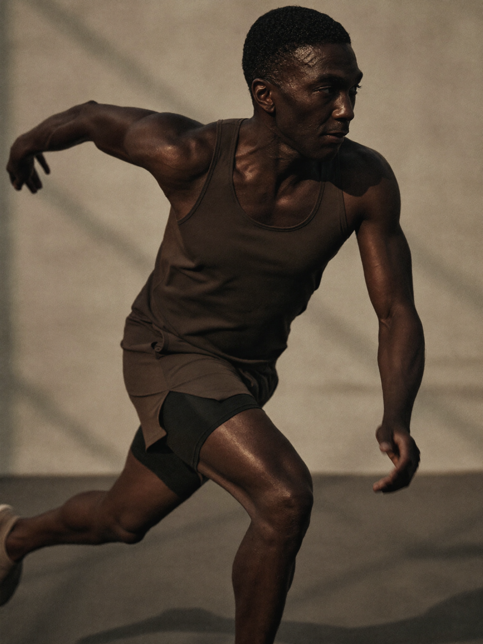 A man running indoors on a smooth, dark floor, wearing a sleeveless athletic shirt and shorts, with his arm extended backward and his head turned to the side.