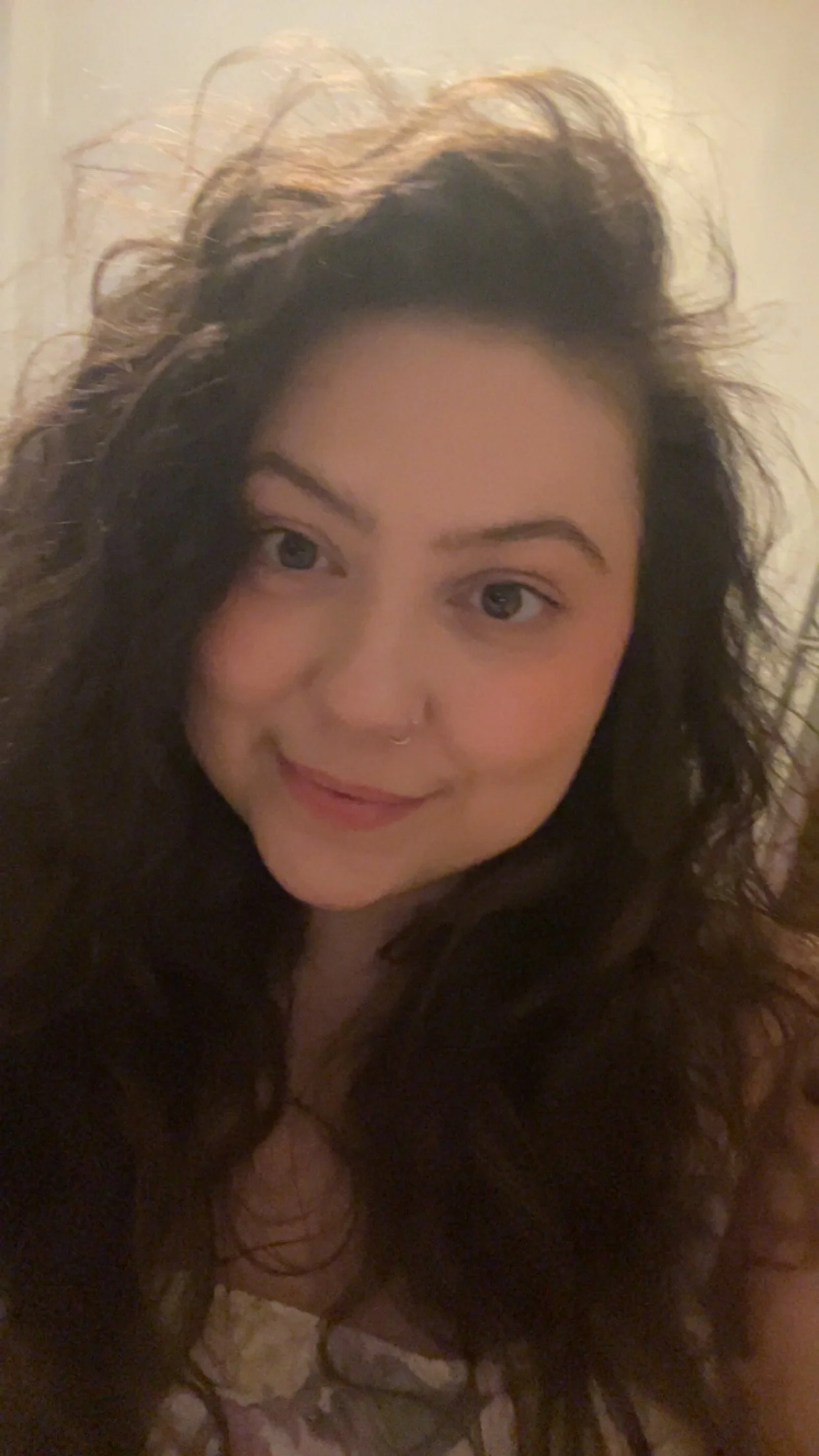 Close-up of a young woman with curly dark hair, smiling, wearing a nose ring and a light-colored top.