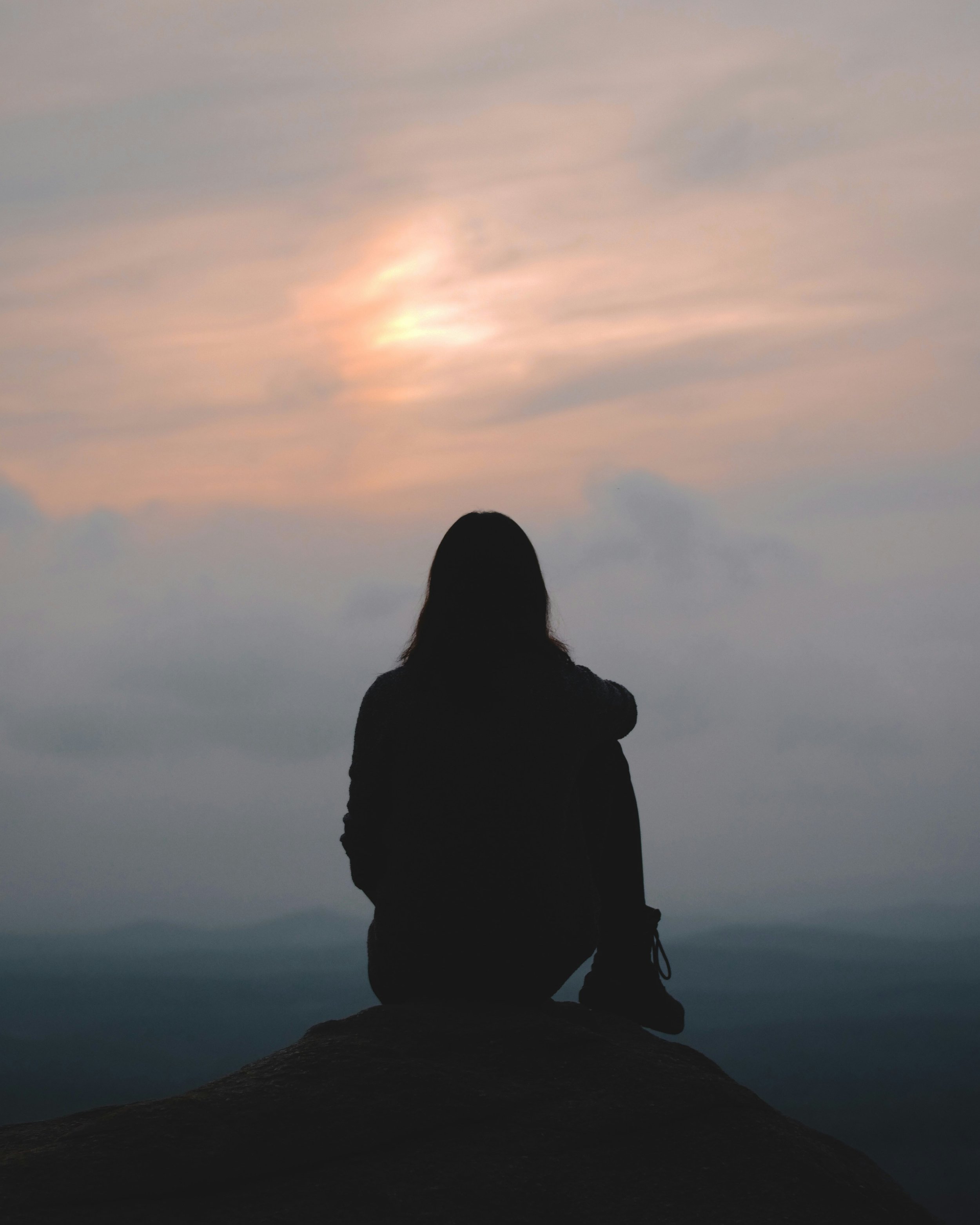 Silhouette of a person sitting on a rock, facing a cloudy sky with a faint sun or moon, during dusk or dawn.