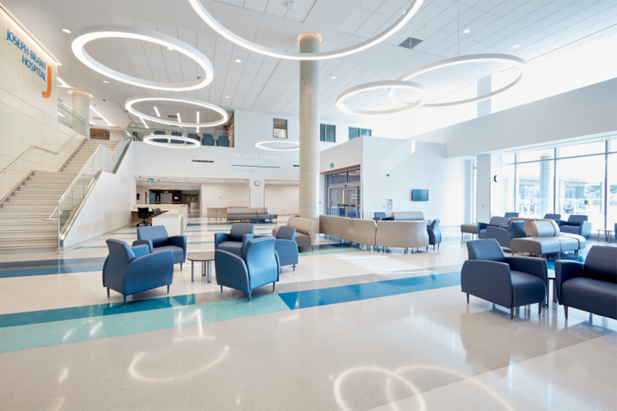 Bright, spacious hospital lobby with blue and beige chairs, a central concrete column, and ceiling lights arranged in circular patterns. Large windows let in natural light. Signage indicates it is Joseph Braumitt Hospital.