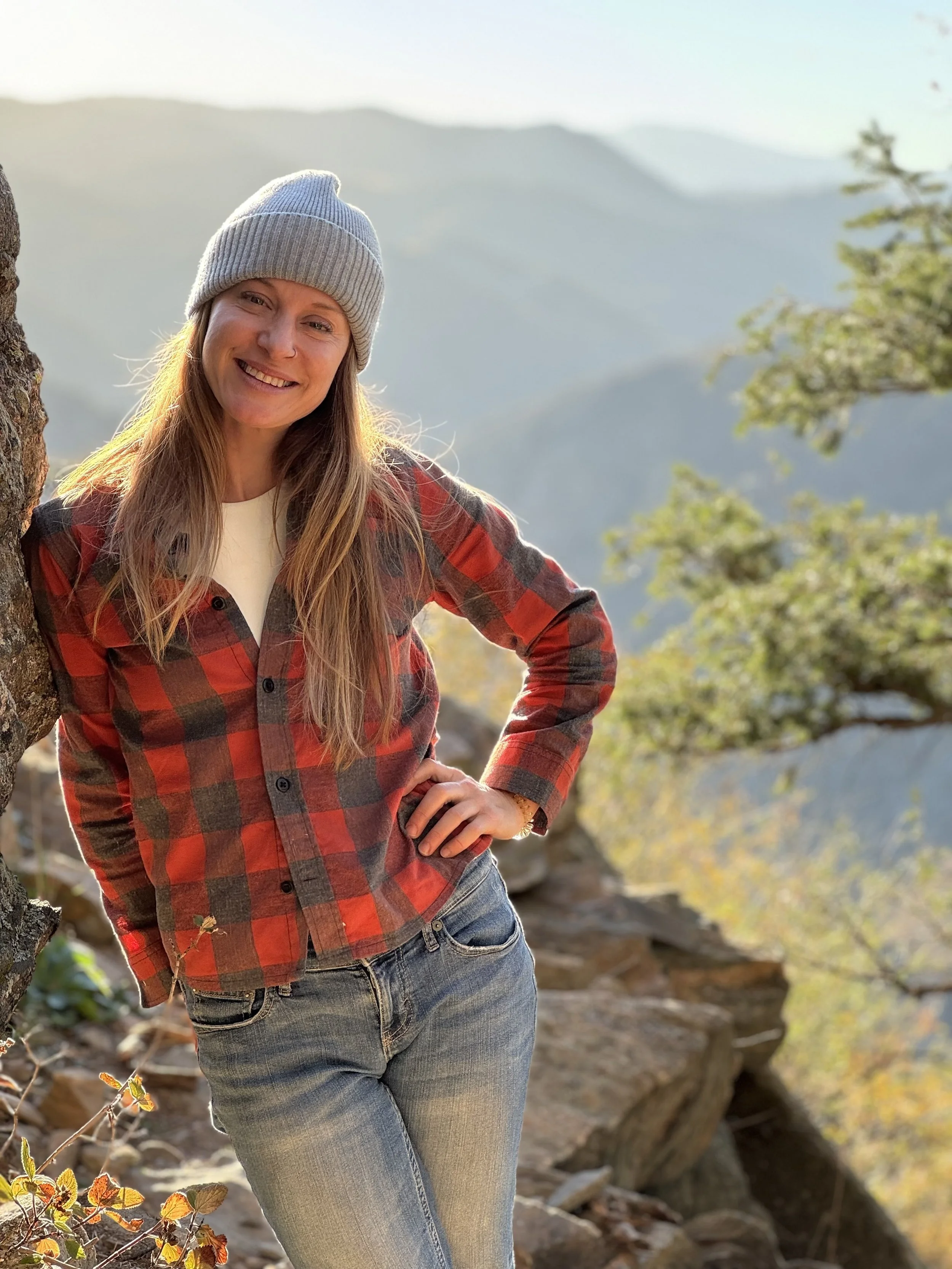 A woman smiling outdoors in a mountainous area, wearing a gray beanie, a red and black plaid shirt, and jeans.