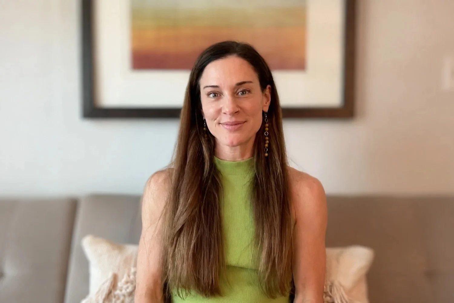 A woman with long brown hair and earrings, wearing a sleeveless lime green top, sitting on a beige couch with pillows, in a room with a framed colorful artwork on the wall behind her.