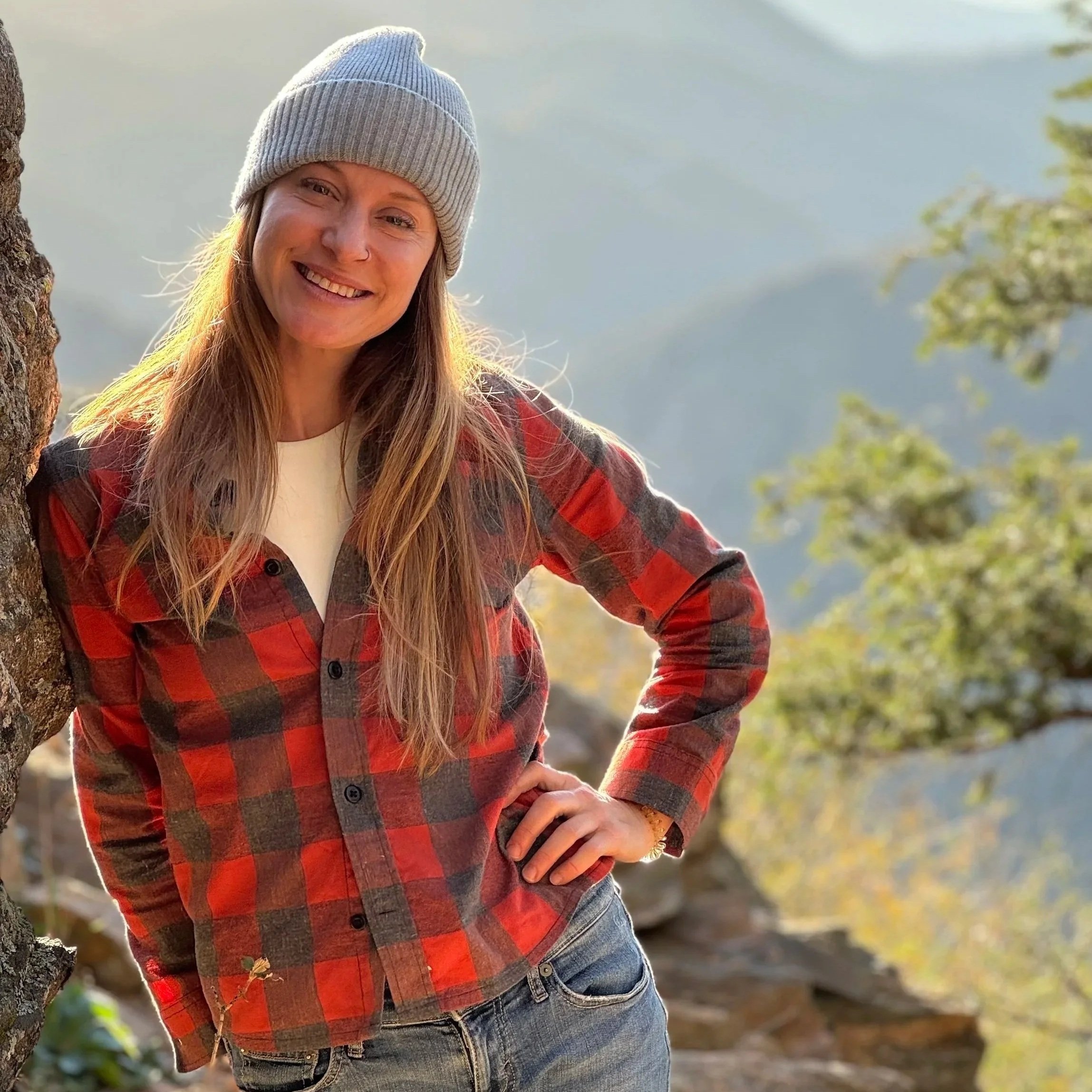 A smiling woman standing outdoors near a tree during daytime, wearing a gray beanie, a red and black plaid shirt, and jeans, with a blurred natural background of water and trees.