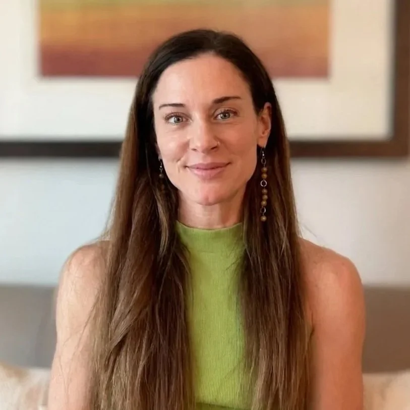 A woman with long brown hair and earrings, wearing a lime green sleeveless top, sitting in a room with a framed artwork in the background.