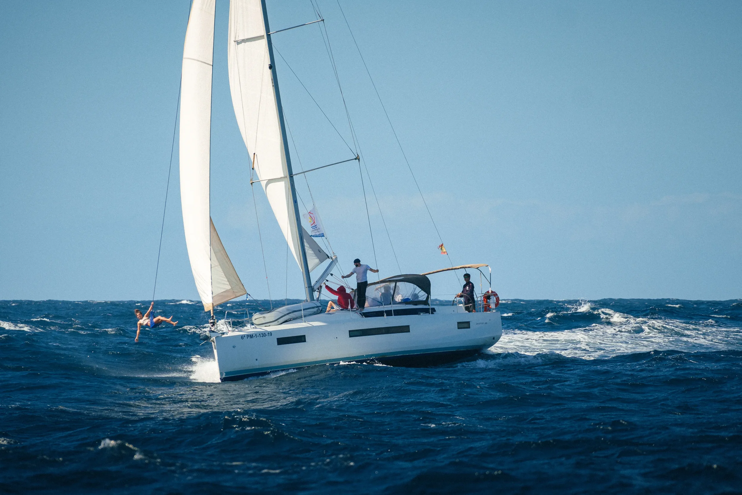 A white sailboat with people onboard sailing on the ocean. One person is hanging off the side in the water, while others are on deck.