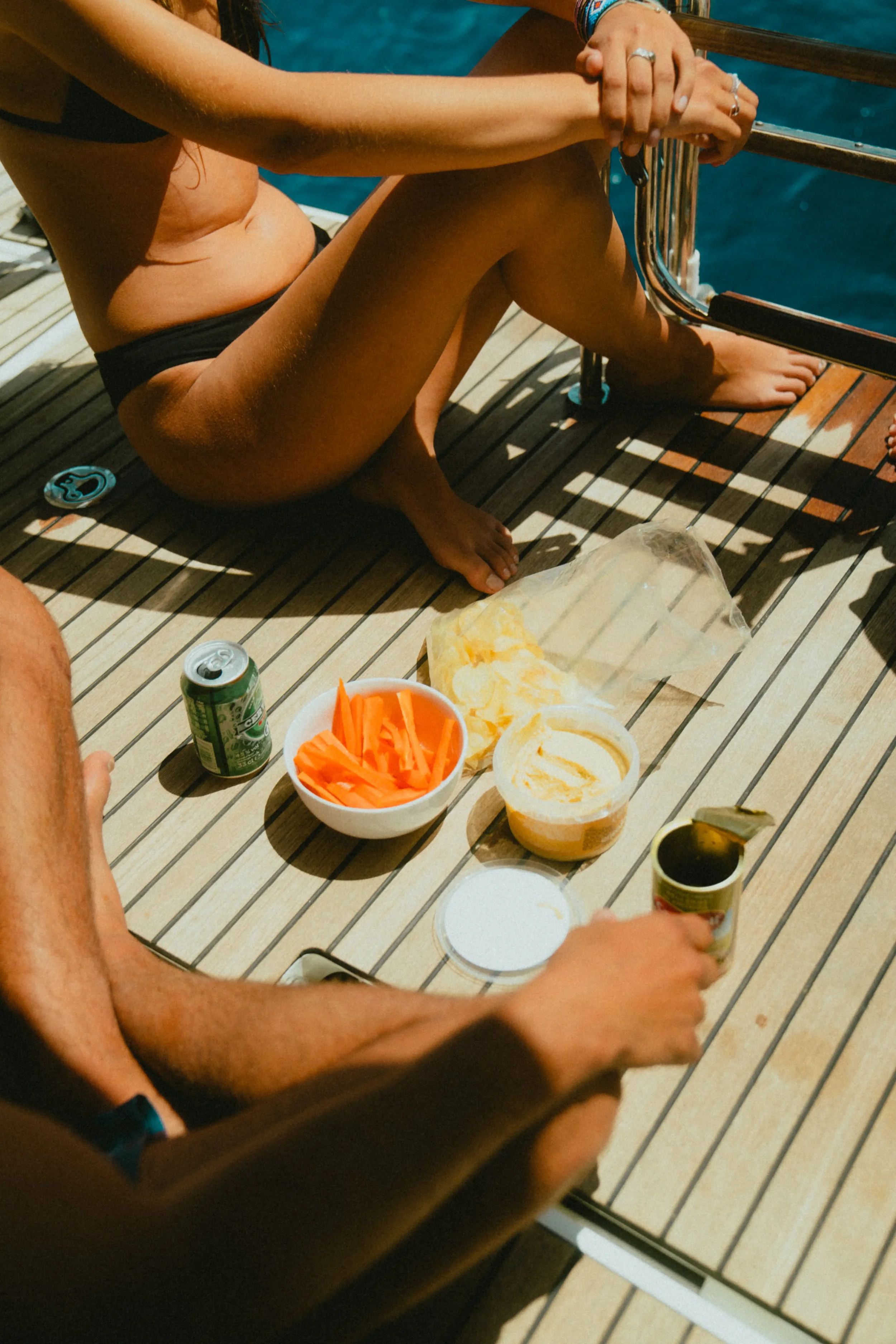 A person sitting on a wooden deck next to water, holding a metal railing. There are snacks and drinks on the deck, including a bowl of sliced carrots, a container with dip, a soda can, and a bottle of wine or champagne. The person is wearing black sh