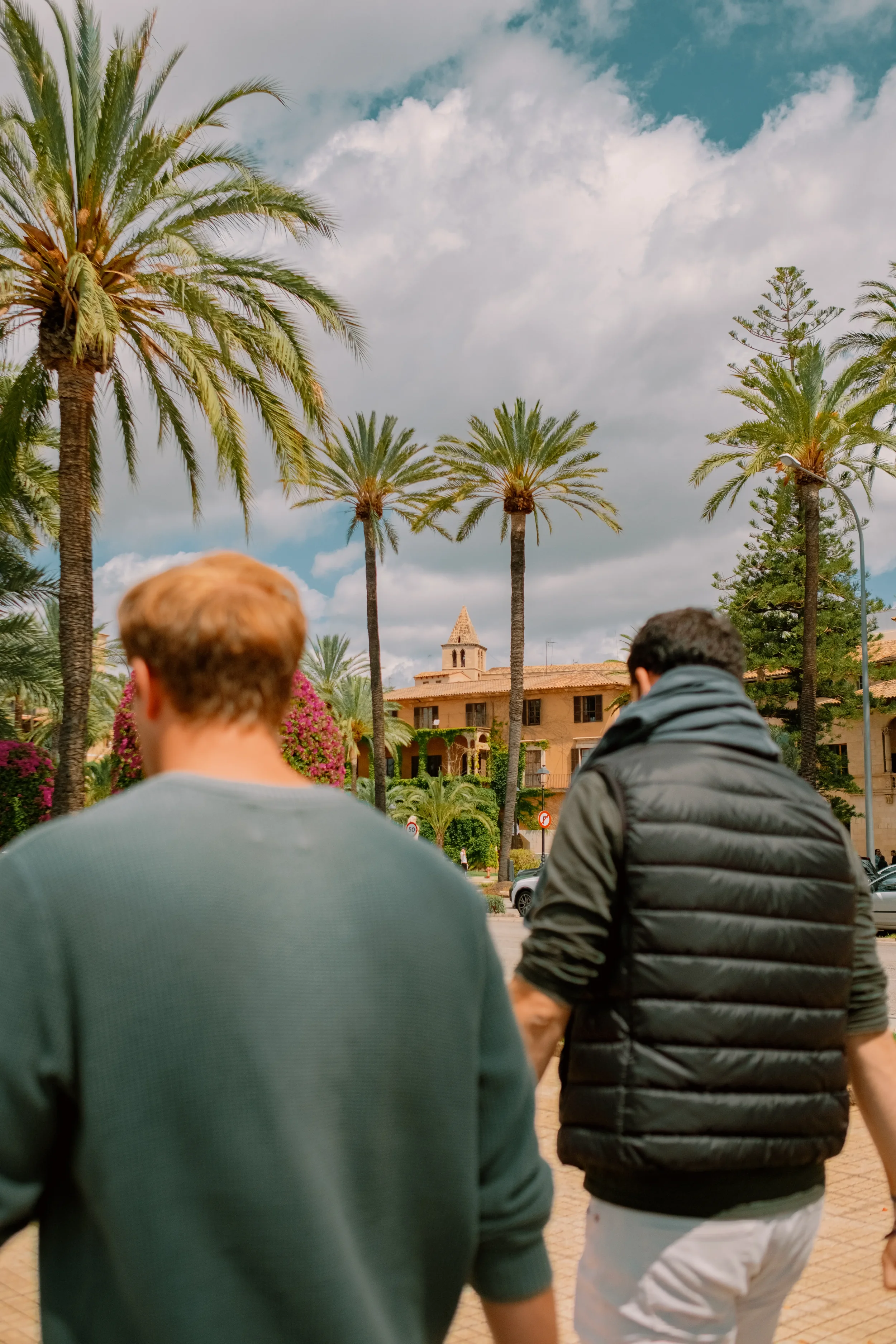 Outside scene with two men walking in front of tall palm trees and a building with a bell tower under a partly cloudy sky.