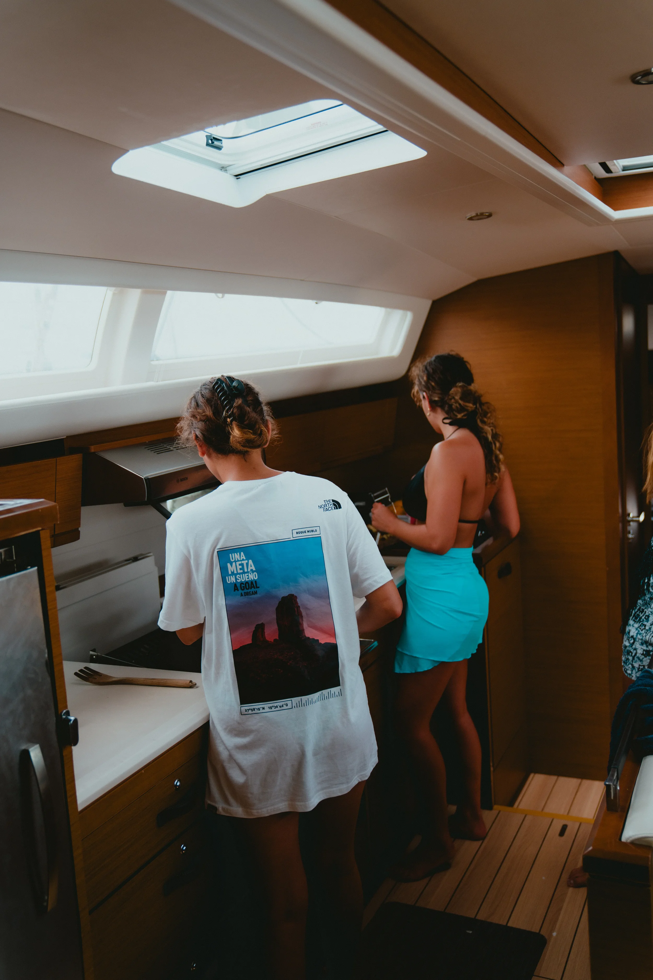 People in a kitchen area on a boat, with one person wearing a graphic t-shirt, another in a black top and blue skirt, and a third partially visible on the right. The ceiling has skylights and the wood-paneled walls create a cozy atmosphere.
