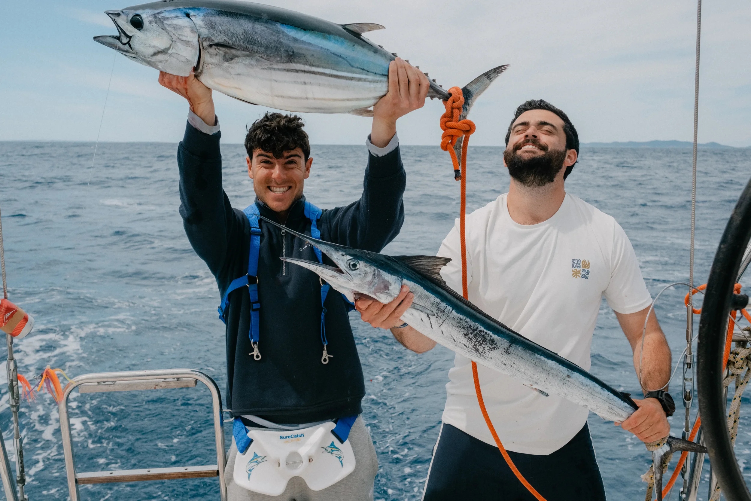Two men on a boat holding a large fish, smiling, with the ocean in the background.