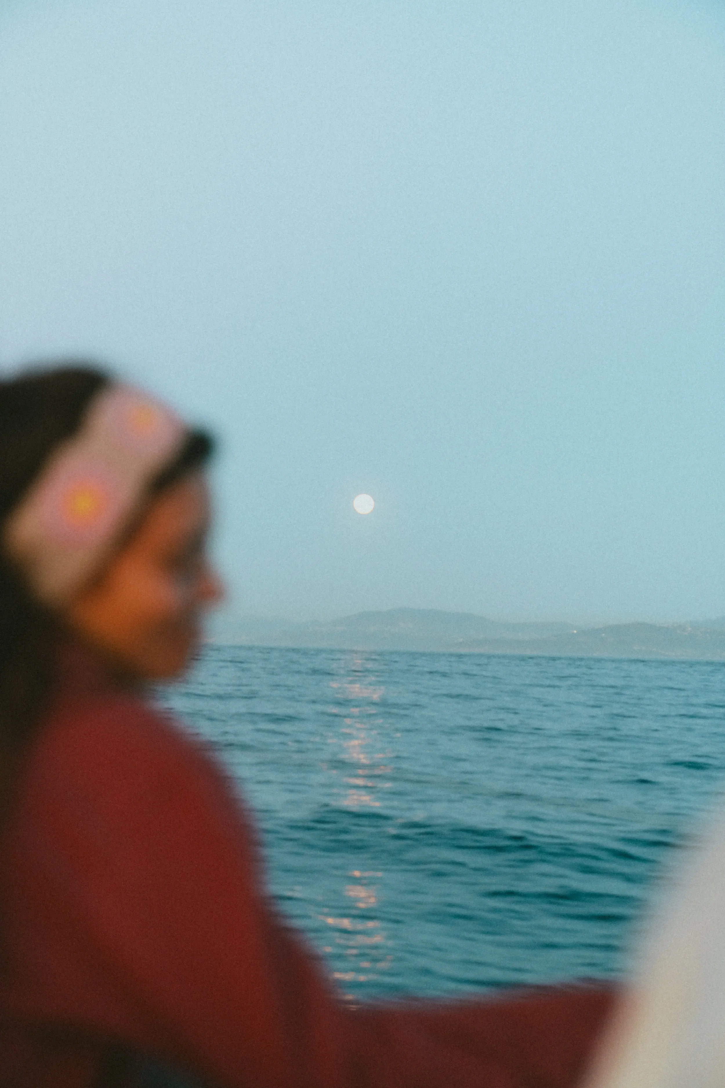 Person looking down, blurred, near a body of water with mountains and a moon or sun in the sky.