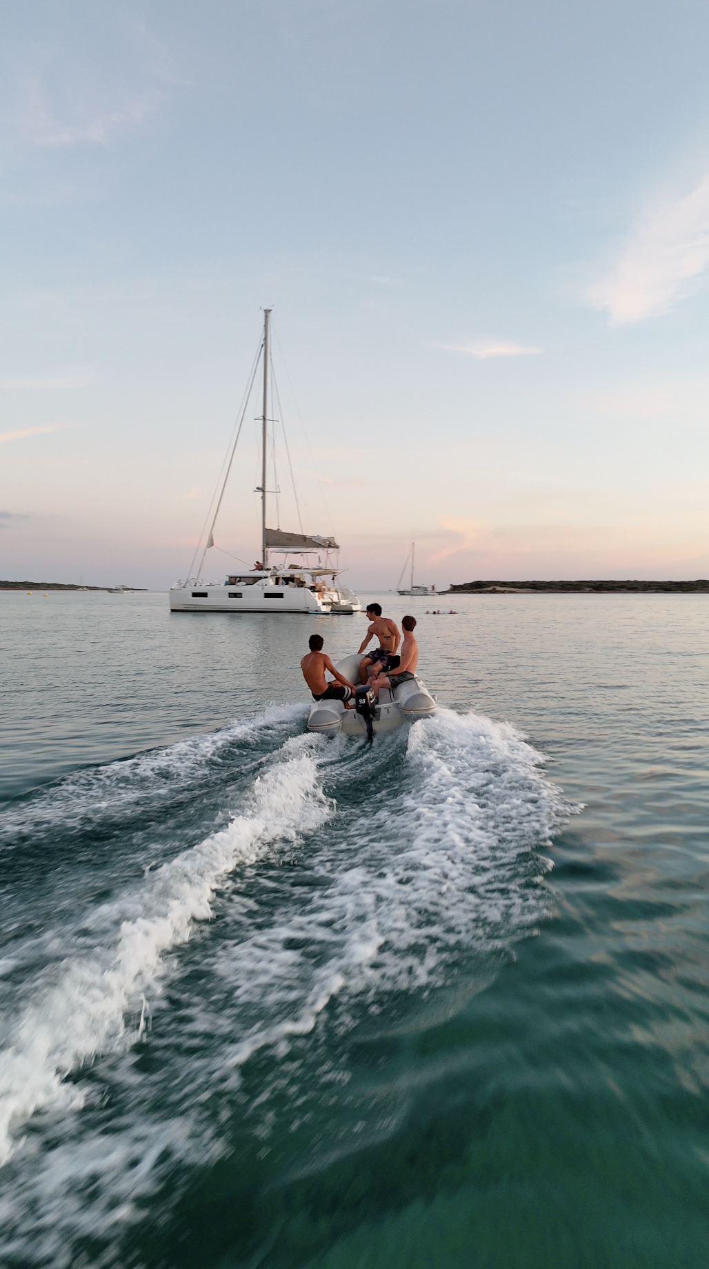 Three young men riding an inflatable boat on a calm body of water near a sailboat at sunset.
