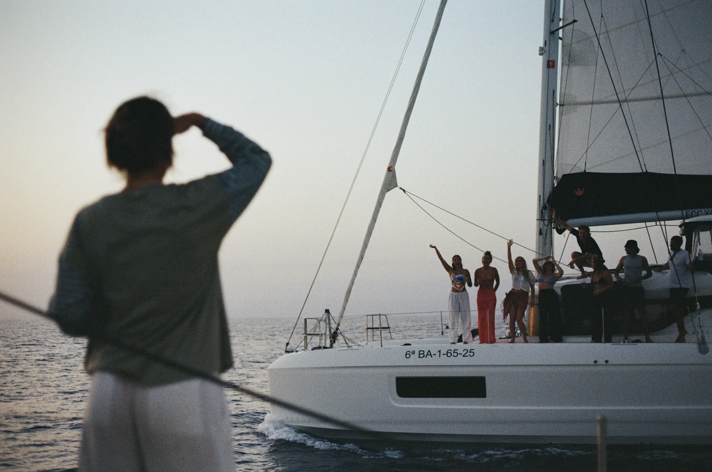 Silhouette of a woman on a boat, with a group of people on the boat deck, and a second woman on the boat deck waving, near the ocean during sunset.