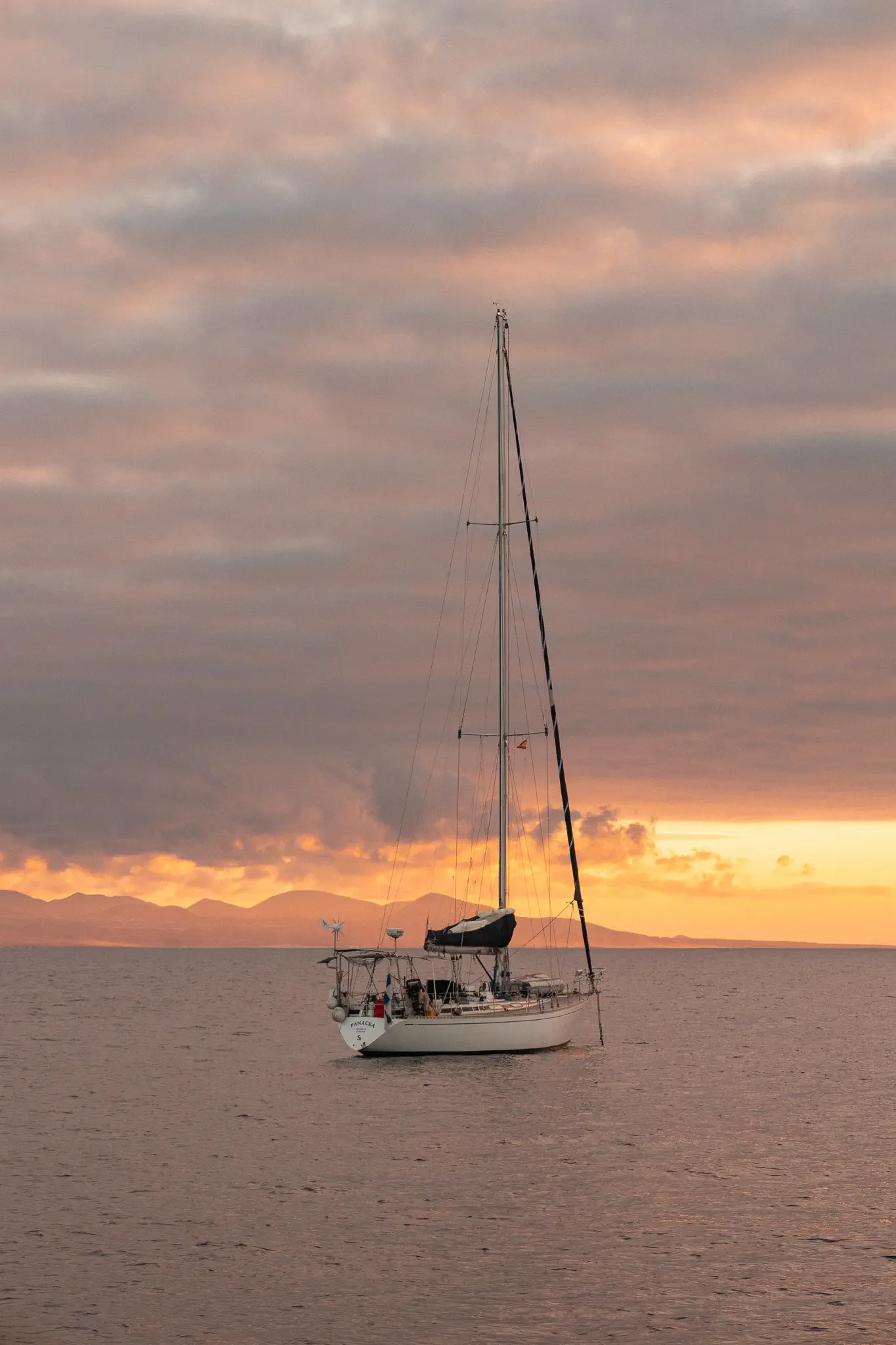 A sailboat on calm water during sunset with a cloudy sky and mountains in the background.