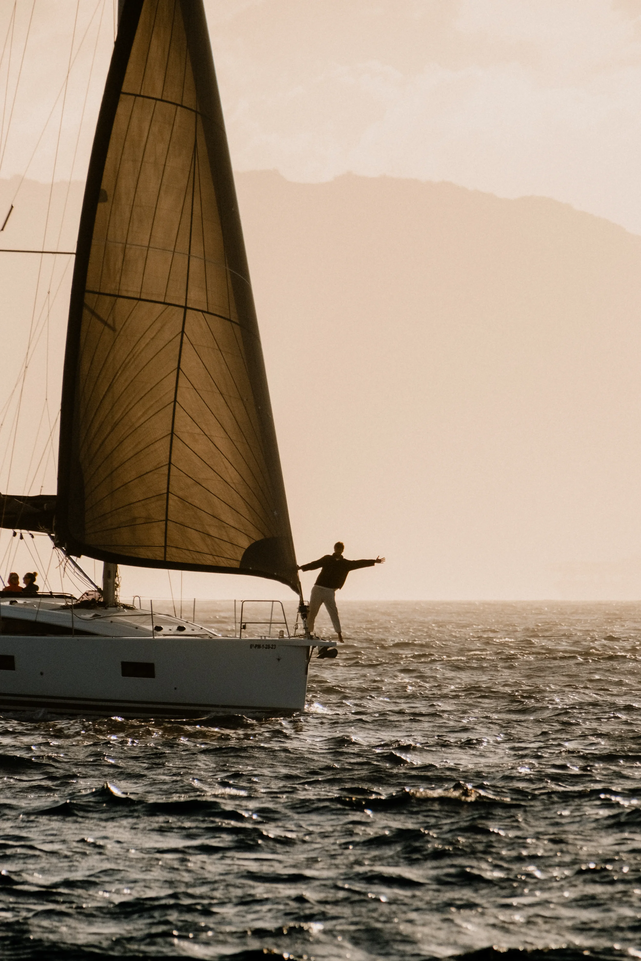 A person standing on the bow of a sailboat with its arms stretched out, sailing on open water during sunset.