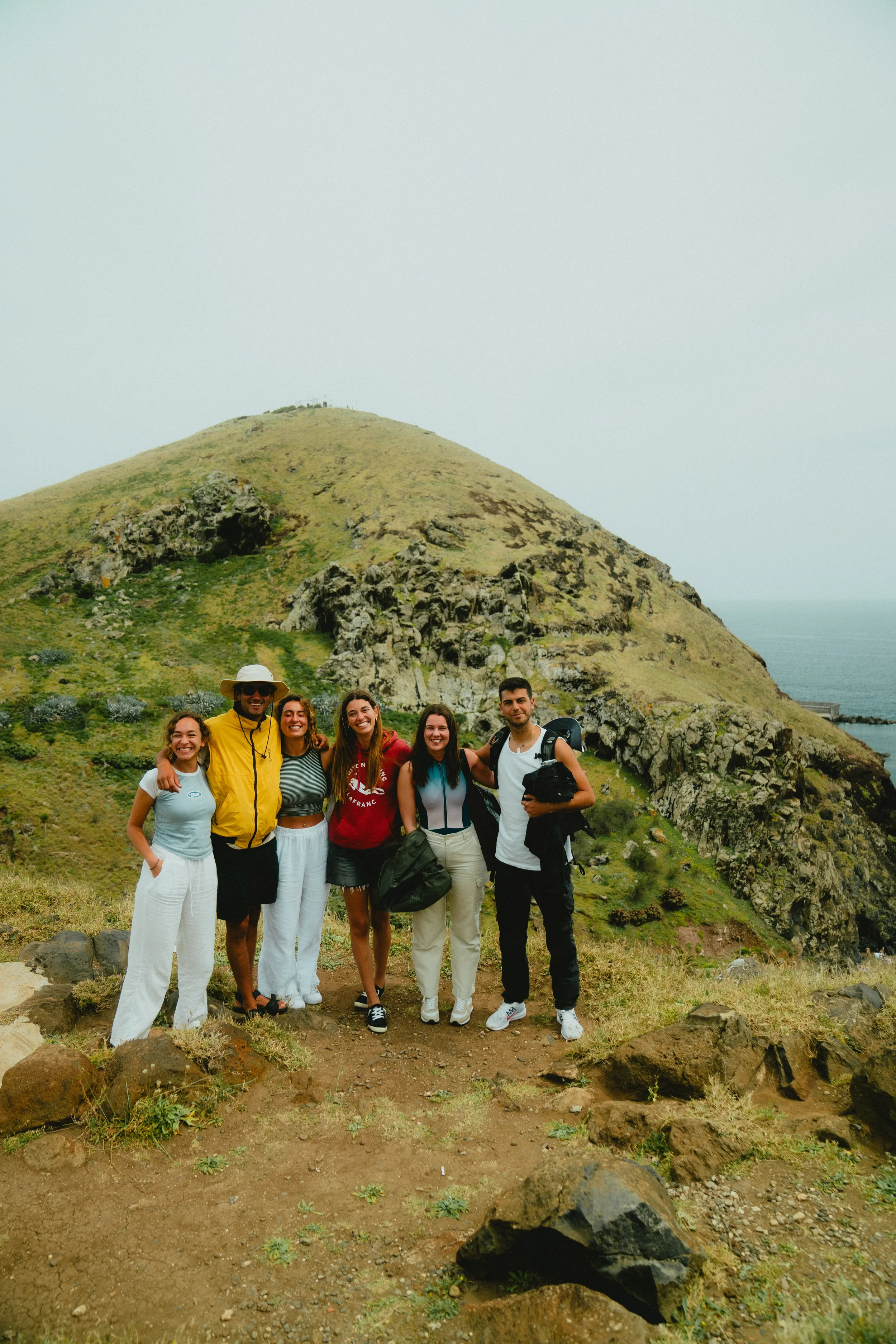 A group of six young people smiling outdoors in front of a grassy hillside with rocks and cliffs, over the ocean on a cloudy day.