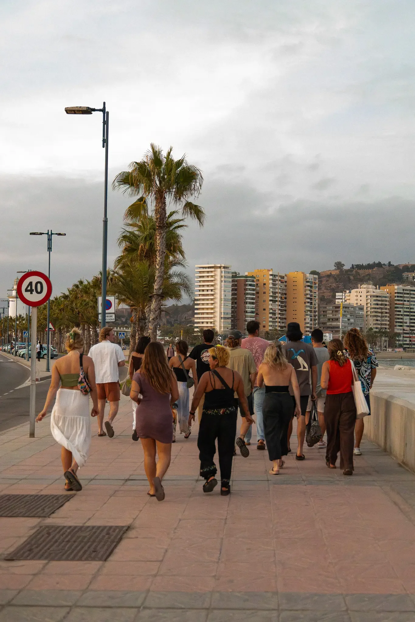 Group of people walking outdoors on a paved promenade with palm trees, high-rise buildings in the background, and dark cloudy sky overhead.