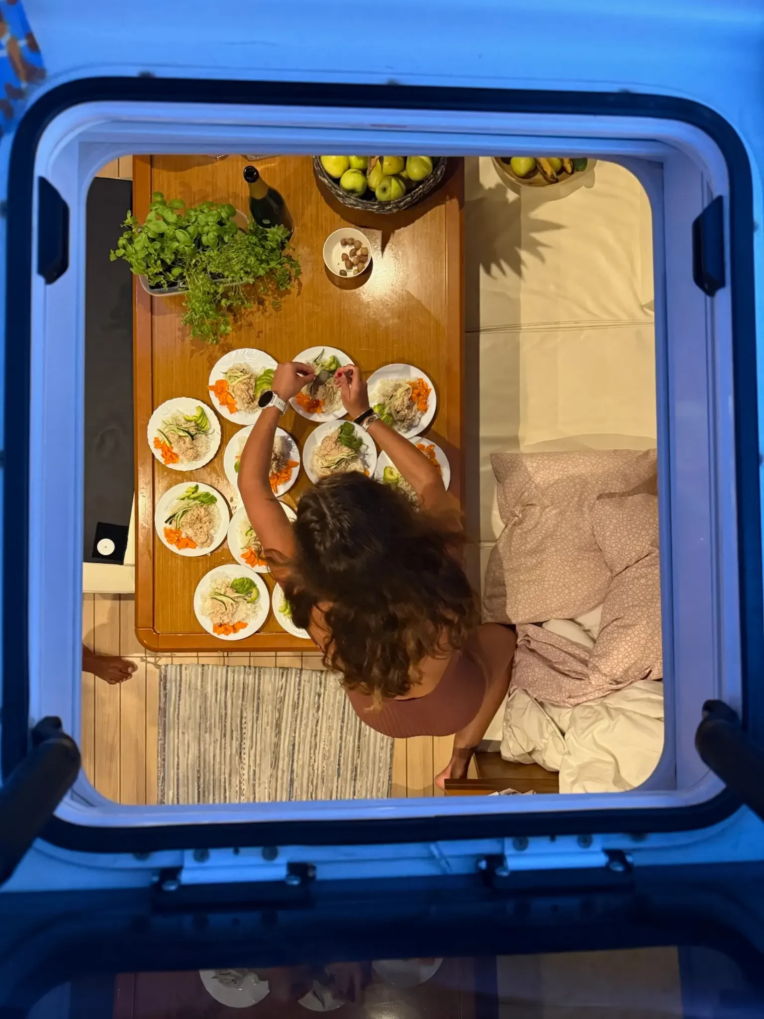 Person preparing plates of food in a kitchen viewed through a blue frame, with a wooden table holding green apples, a bottle, and a bowl of nuts.