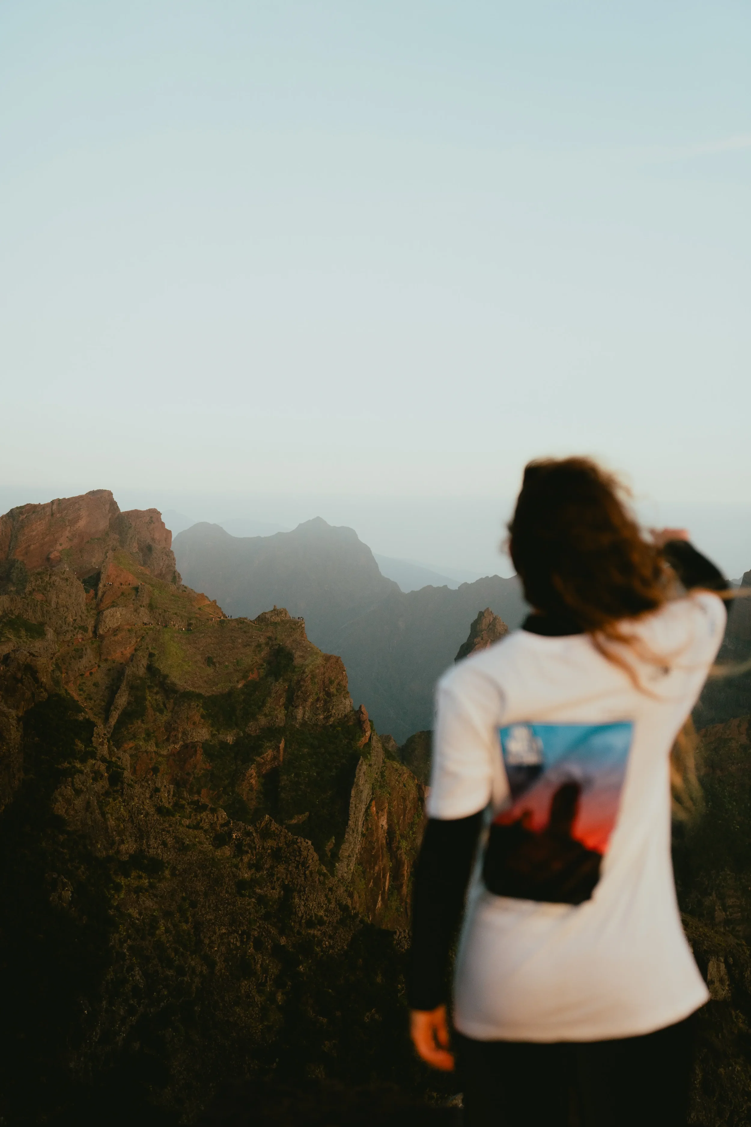 A person standing on a rocky mountain overlooking a mountain range as the sun sets or rises in the sky.