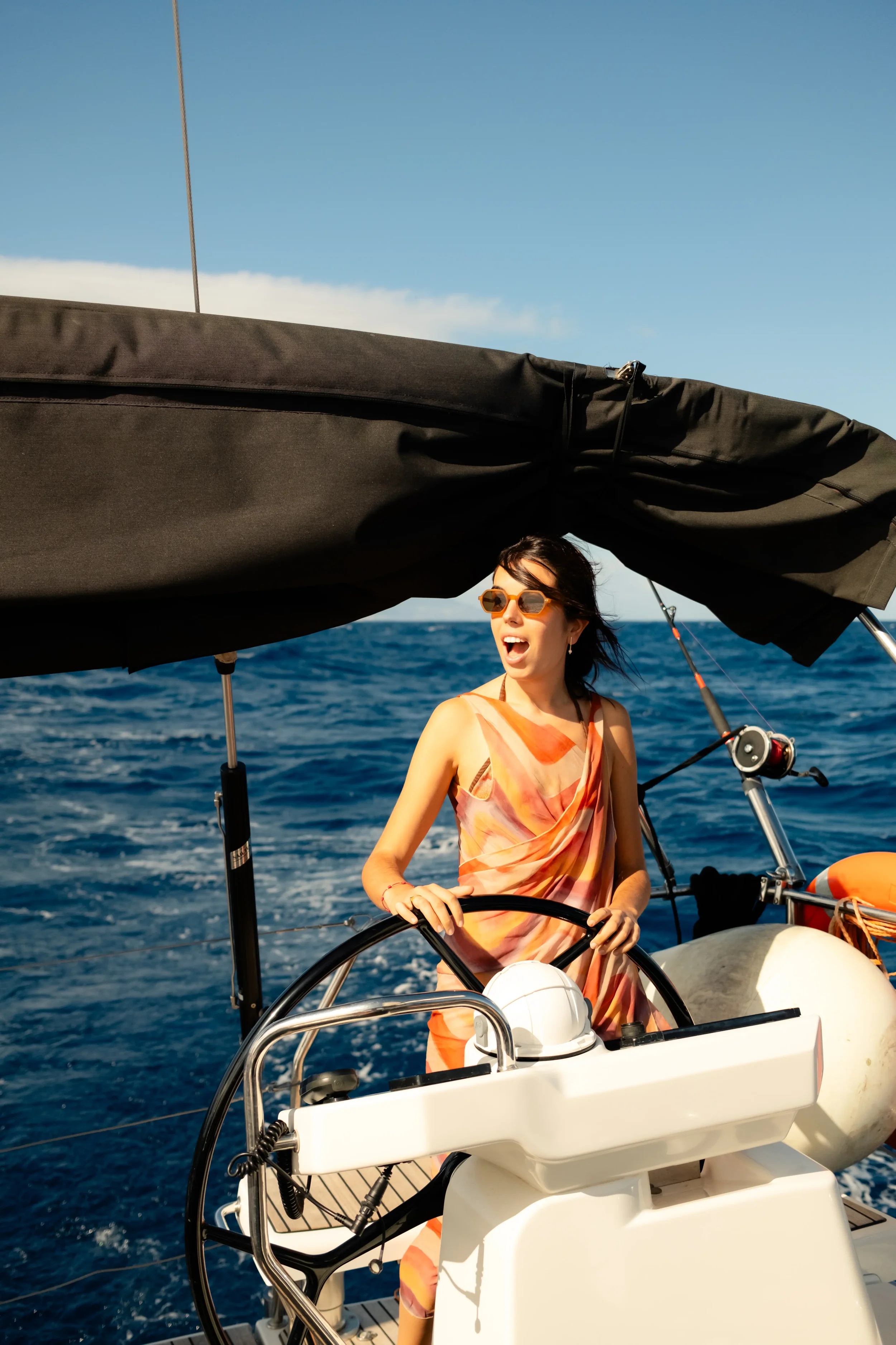 Woman wearing sunglasses and a colorful dress sailing a boat on the ocean, with a blue sky and a few clouds in the background.