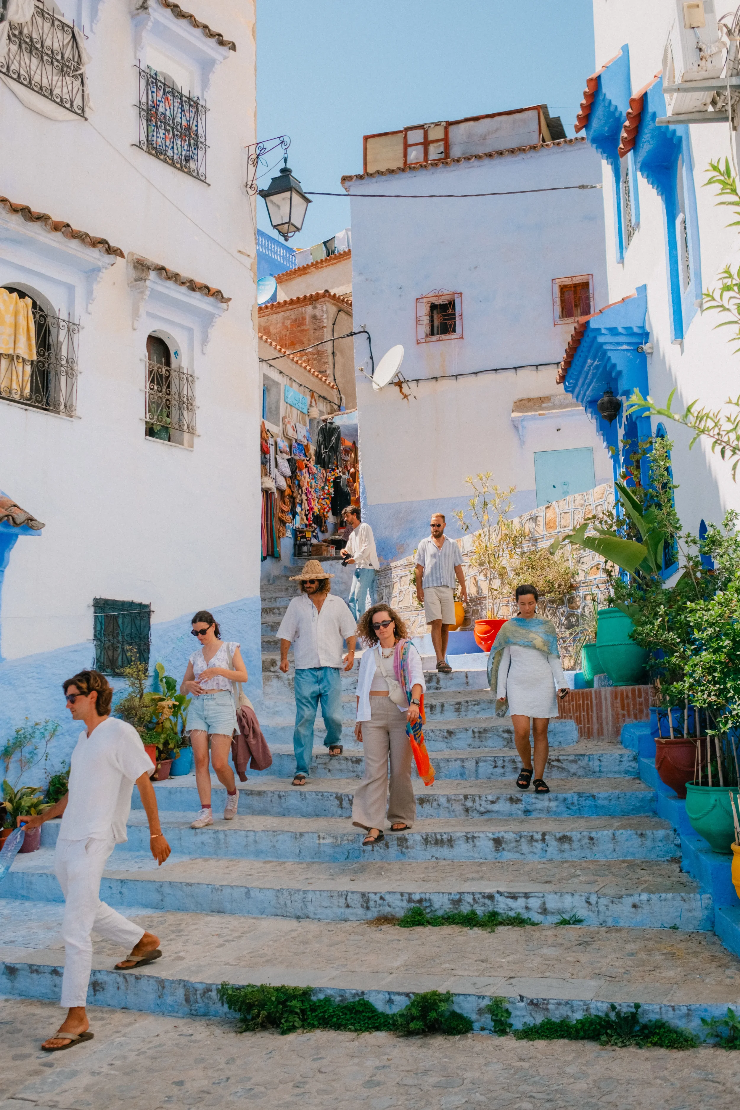People walking down outdoor stairs in a colorful, Mediterranean-style town with white and blue buildings, laundry hanging, and potted plants.