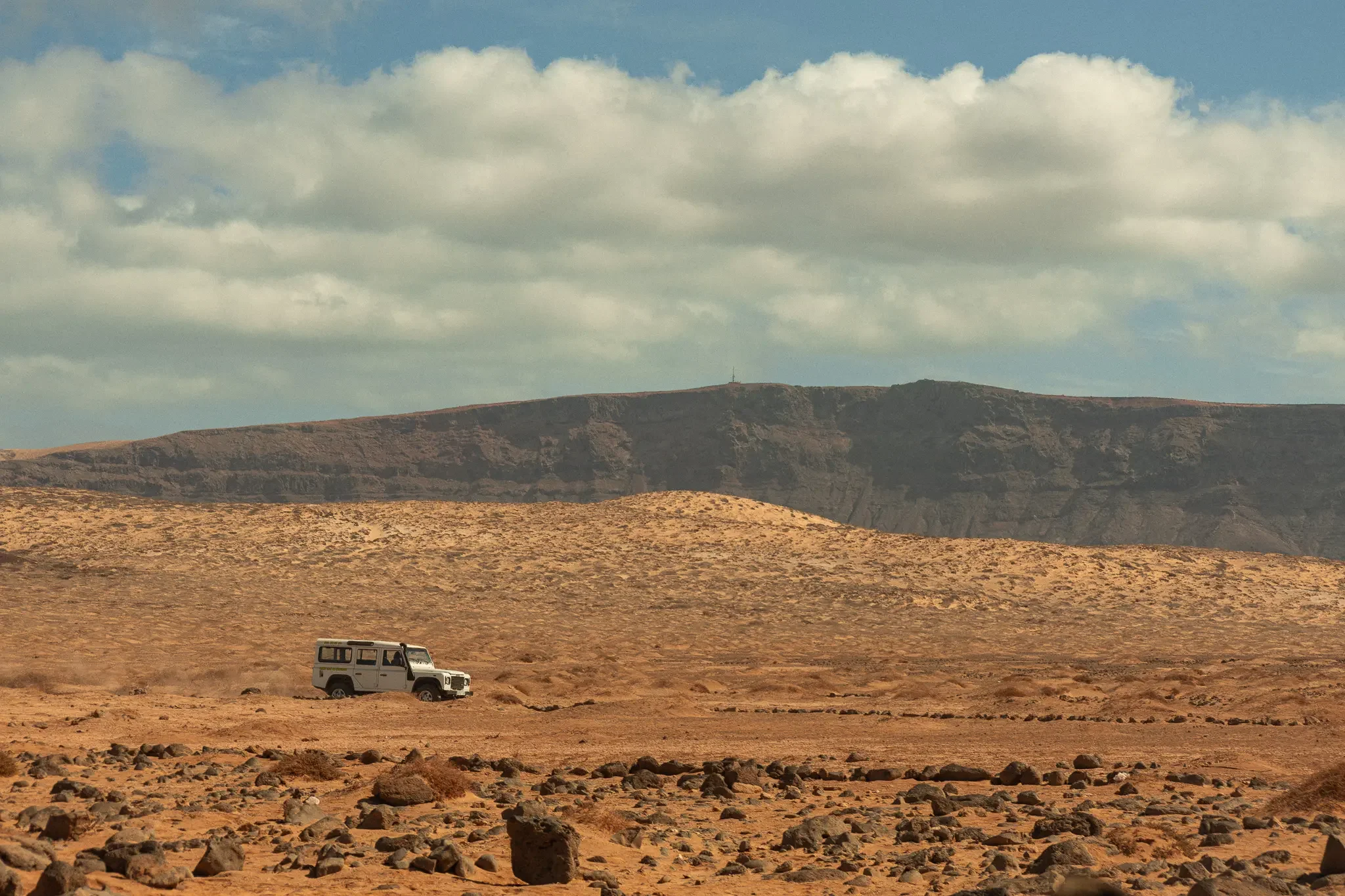 A white off-road vehicle parked on a barren desert landscape with rocky terrain and mesas in the background under a partly cloudy sky.