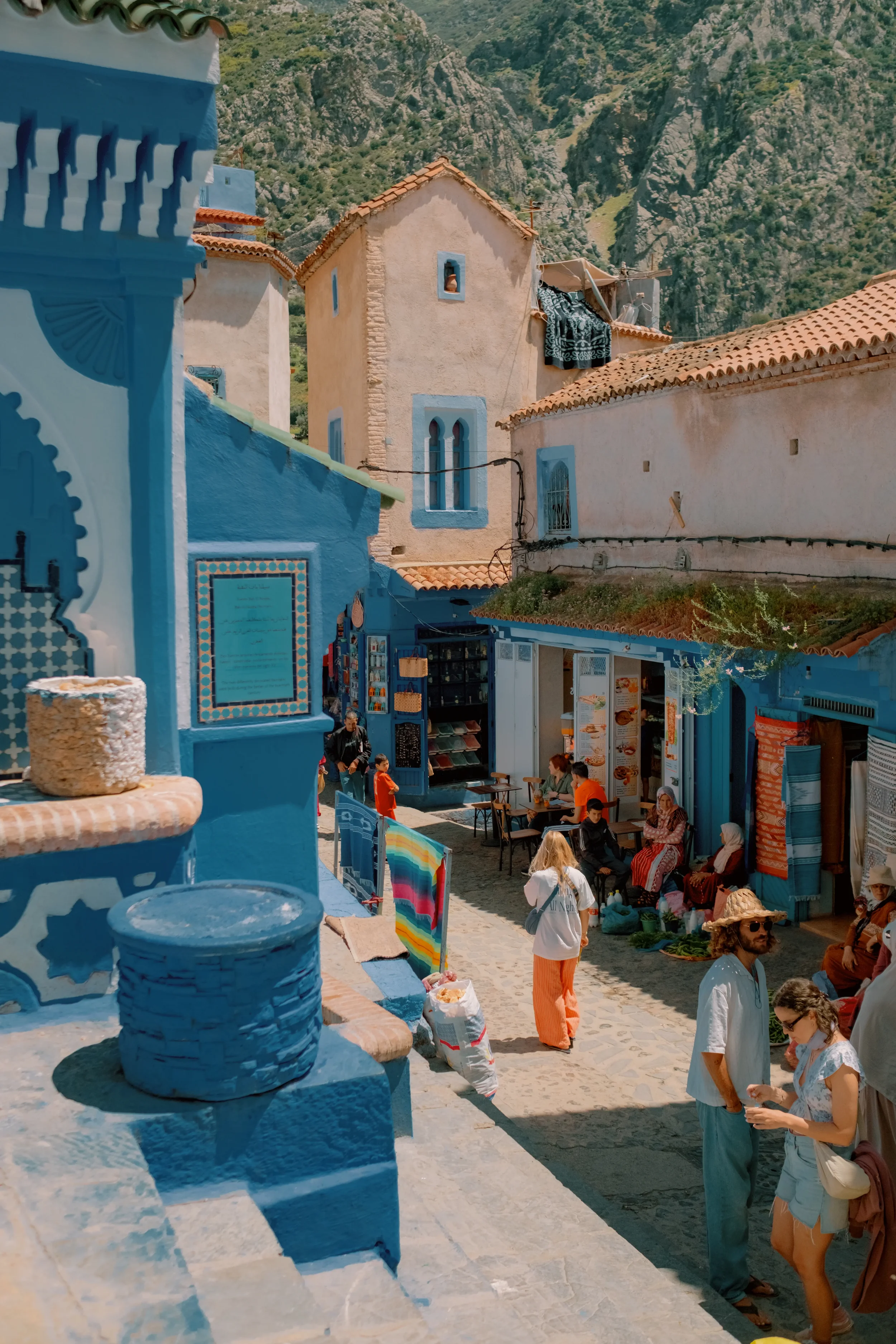 Scene of a street in a town with buildings painted in blue and white, people walking and browsing shops, mountainous landscape in the background.