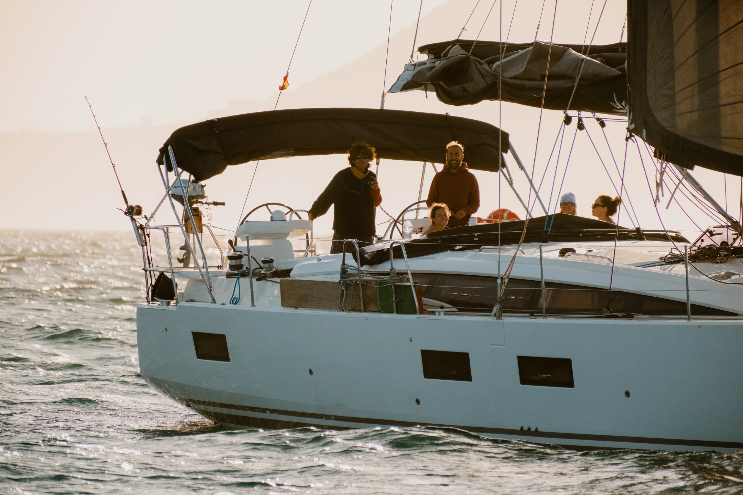 Two men and two women sailing on a boat with a black canopy, with the sun setting or rising in the background over the water.
