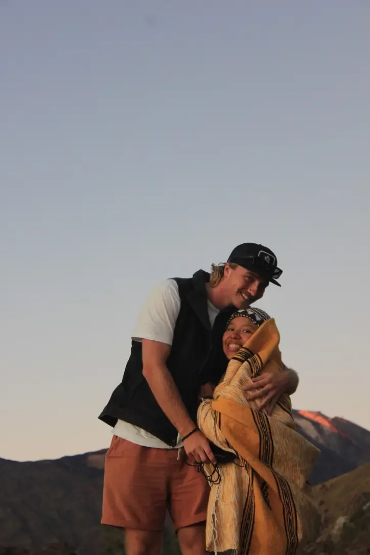 A man and a woman embracing outdoors with mountains in the background, during sunset or twilight.