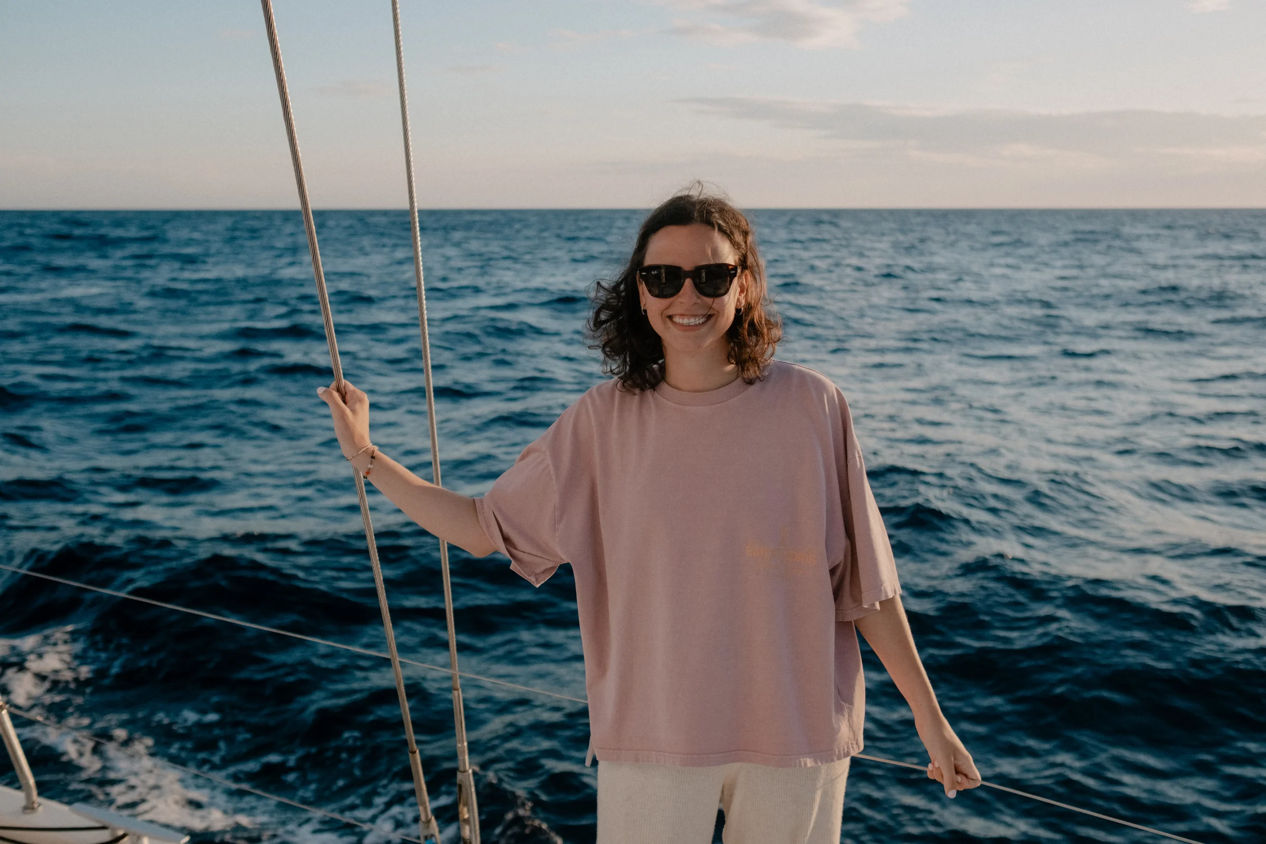 A woman with curly hair wearing sunglasses and a pink t-shirt smiling on a boat, holding a rope, with the ocean and sky in the background.