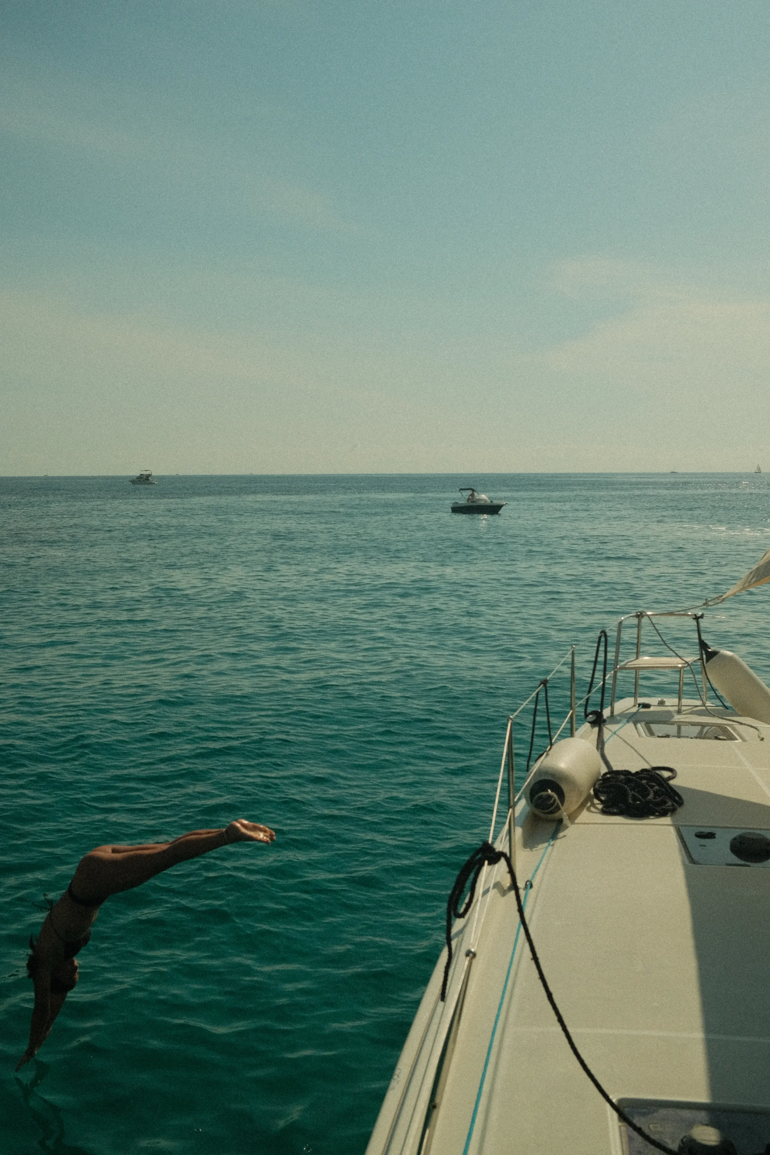 A person is diving into the ocean from a boat. The boat has various ropes and equipment on its deck. Several boats are visible in the distance on the calm water, under a clear sky.