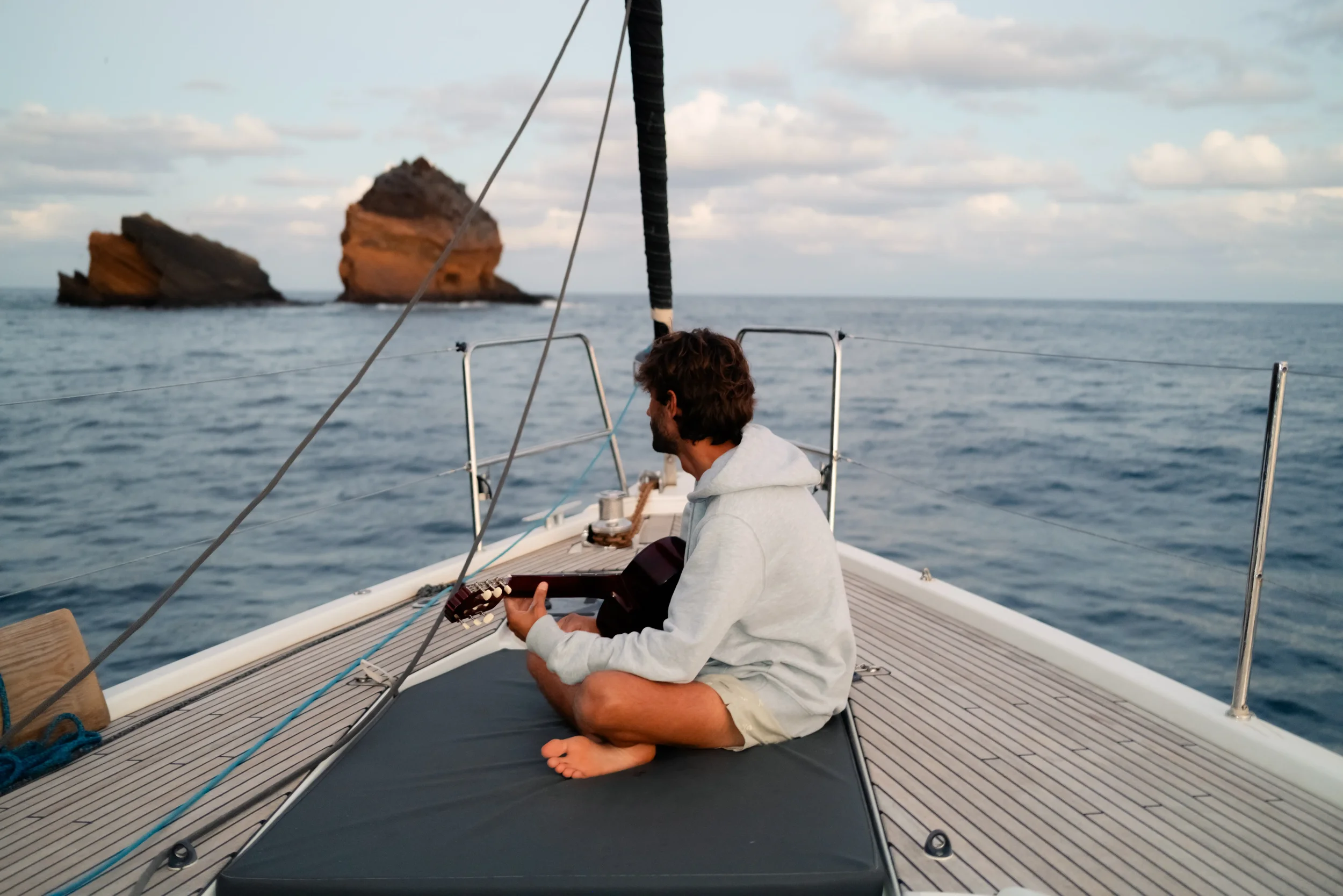 A person sitting barefoot on a boat, holding a guitar, with two large rocky islands in the distance and a partly cloudy sky.
