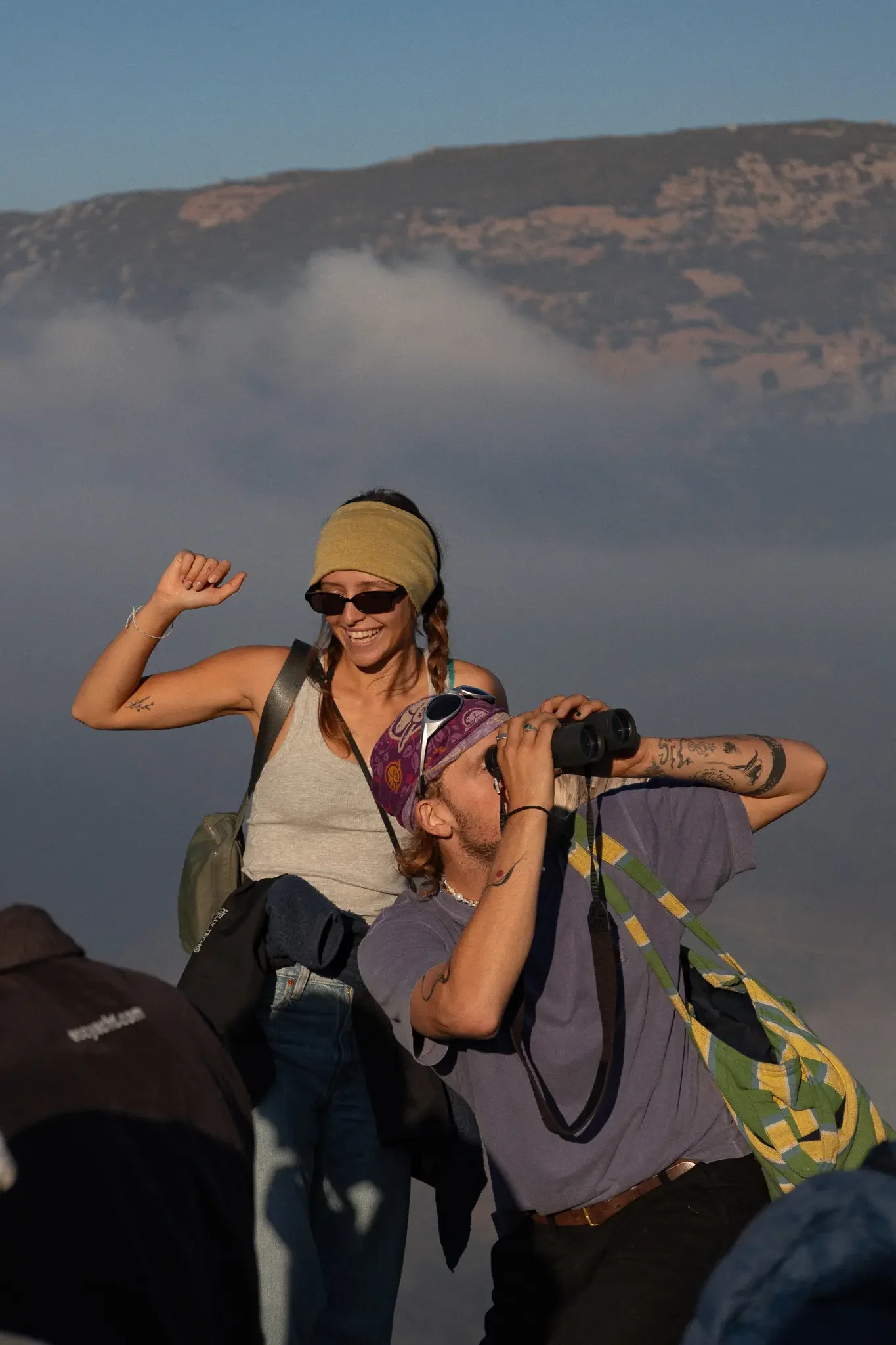 Two women and a man with backpacks are in a foggy outdoor setting with hills in the background. The woman in the foreground wears sunglasses, a headband, and a tank top. The woman behind her has braided hair, sunglasses, and a tank top, smiling and r