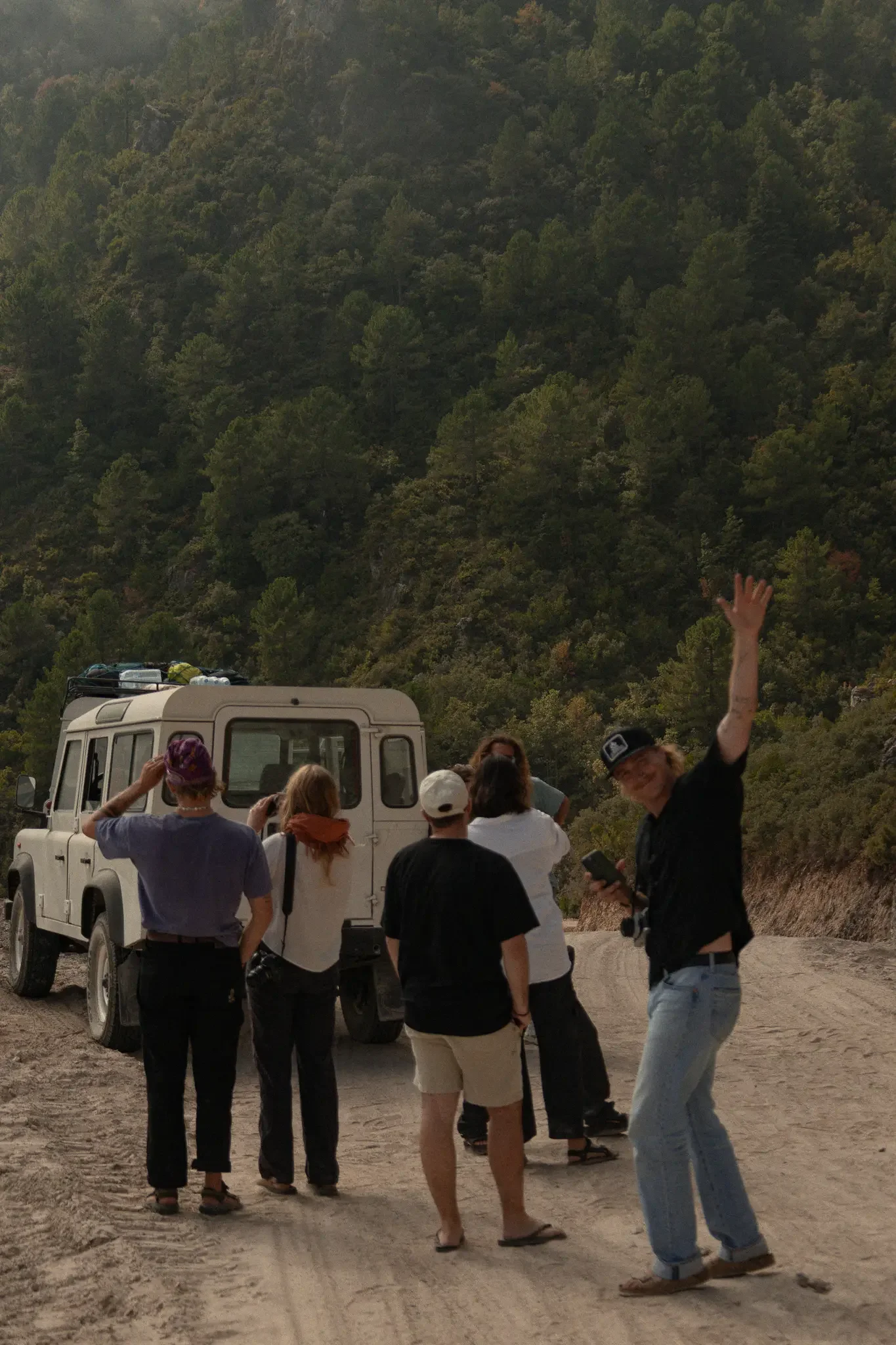 A group of people with cameras and phones standing on a dirt road in front of a white jeep, with a forested hillside in the background, some waving or looking at the camera.
