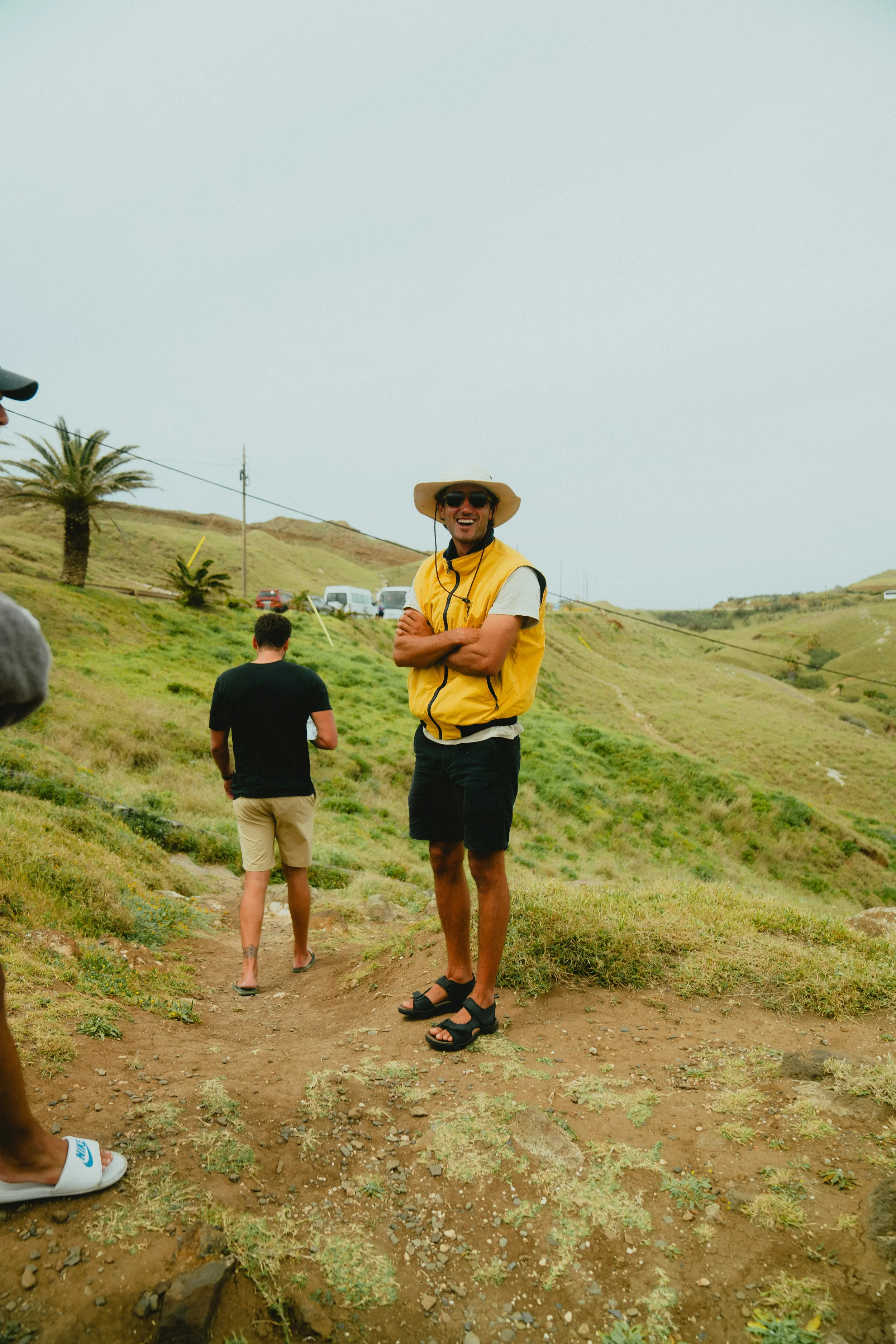 Smiling young man in a yellow vest, wide-brimmed hat, sunglasses, and sandals standing on a dirt path in a grassy hillside area with other people nearby.