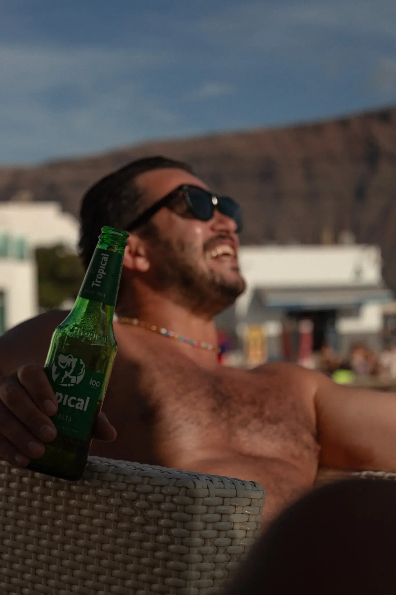 A shirtless man wearing sunglasses laughing and holding a green bottle of Topcical beer in his hand, sitting next to a woven basket, with a mountain and sky in the background.