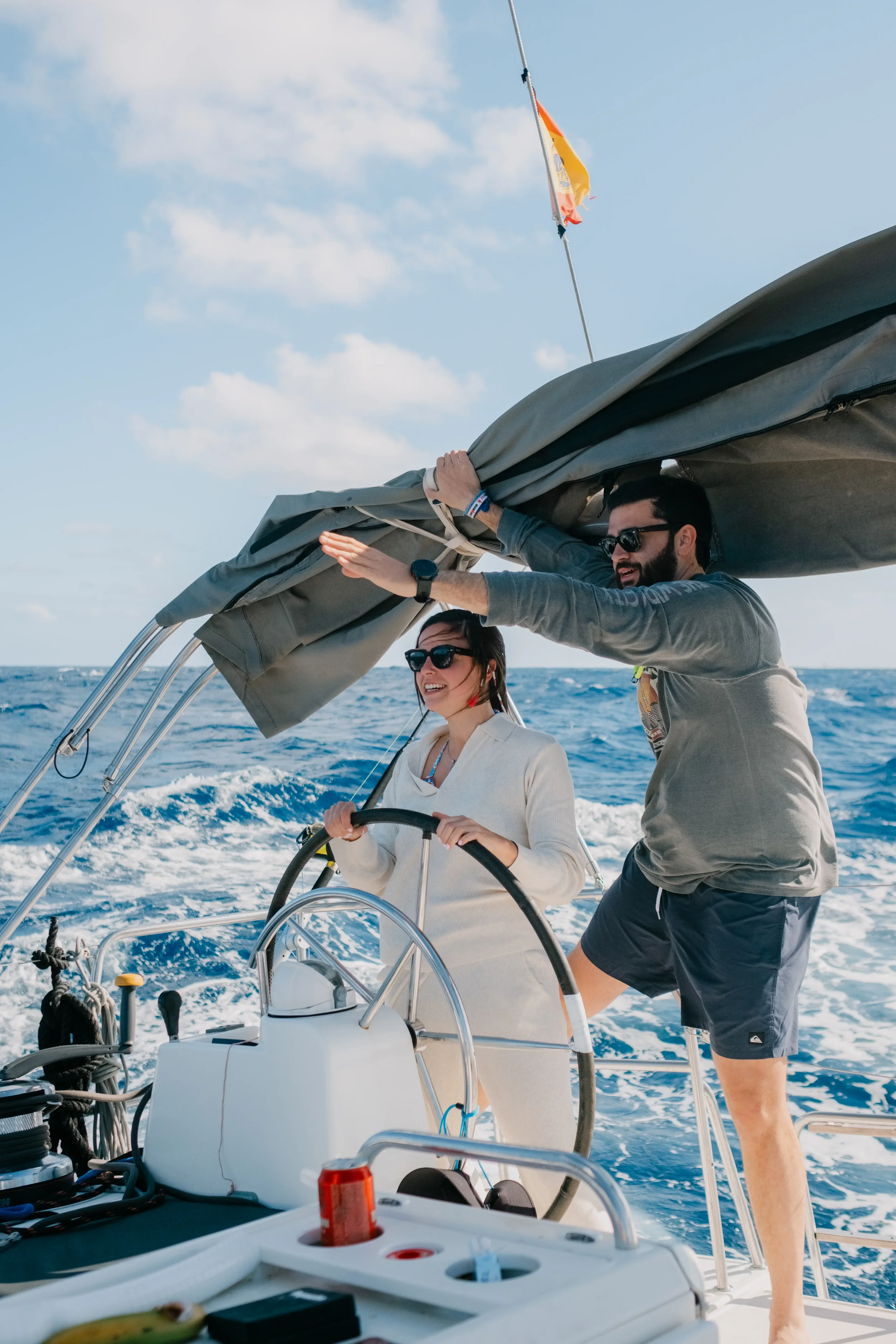 Two people sailing on a boat in the ocean; one man and one woman, steering the boat and enjoying the moment with clear blue sky and some clouds in the background.