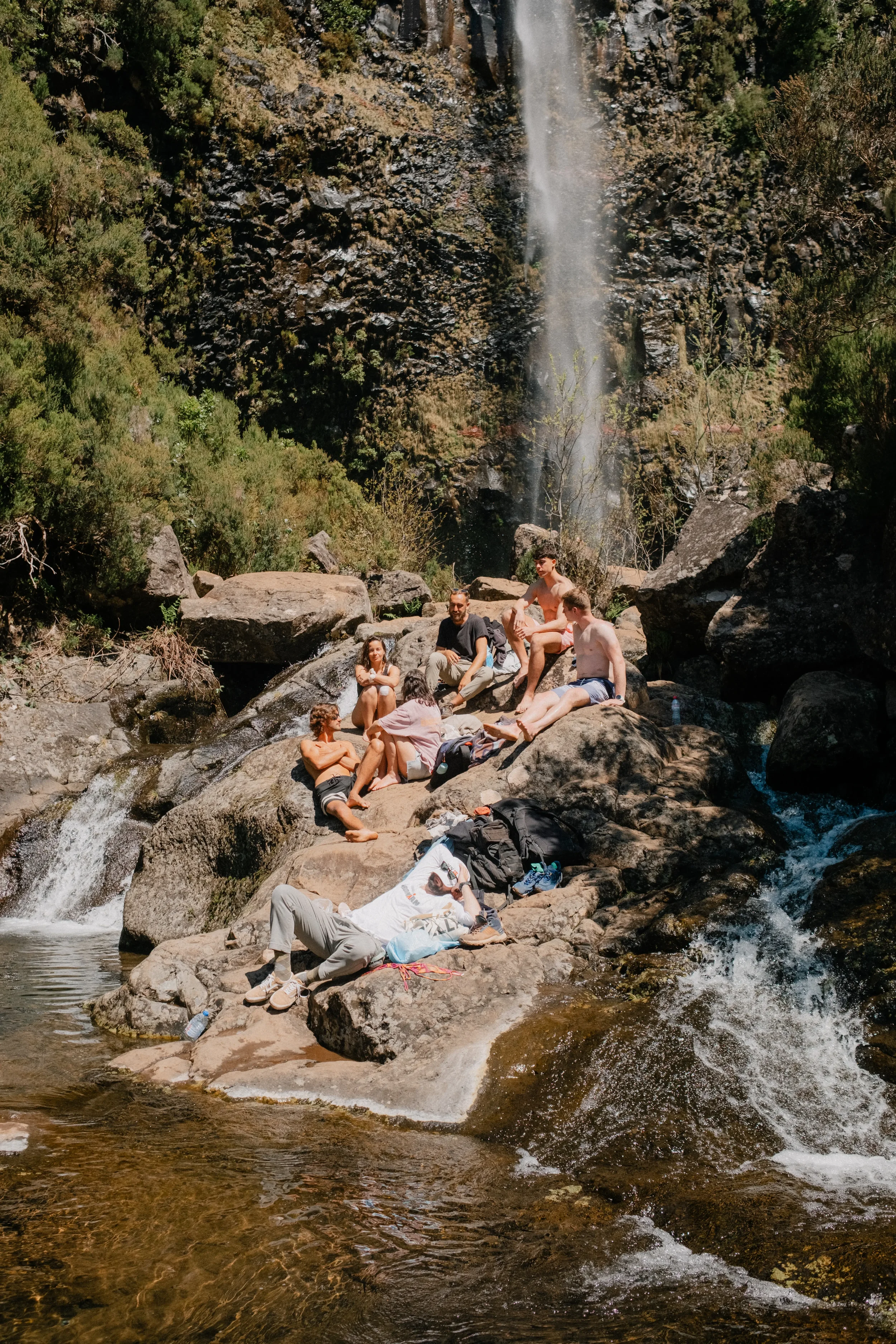 Group of people relaxing on rocks in a stream near a waterfall, surrounded by greenery and trees in a natural outdoor setting.