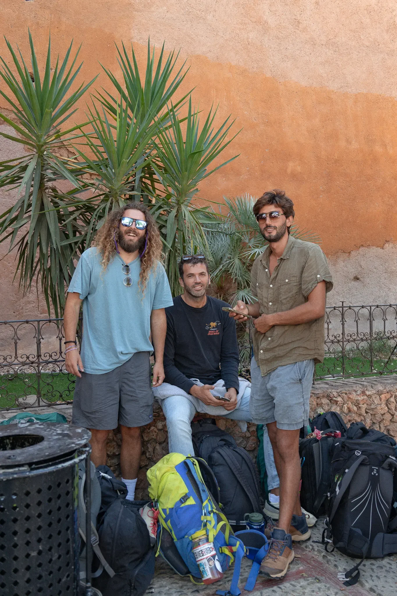 A group of three men outdoors in front of a large, orange concrete wall with desert plants behind them. They are dressed casually, with backpacks and hiking gear around them, and are smiling while holding backpacks and phones.