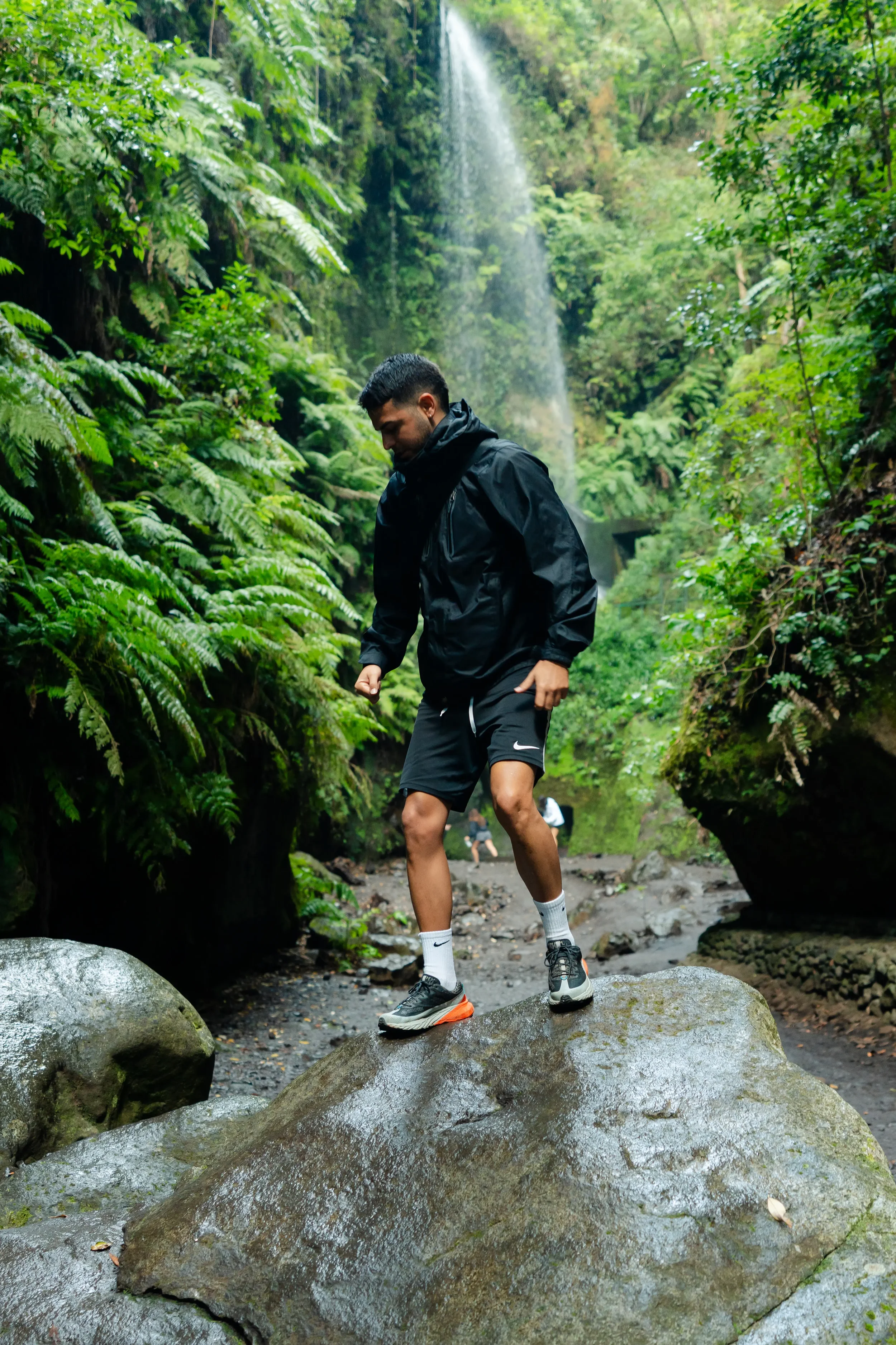 A man in a black rain jacket and black shorts stands on a large wet rock in a lush green forest with ferns and trees. A waterfall is visible in the background.
